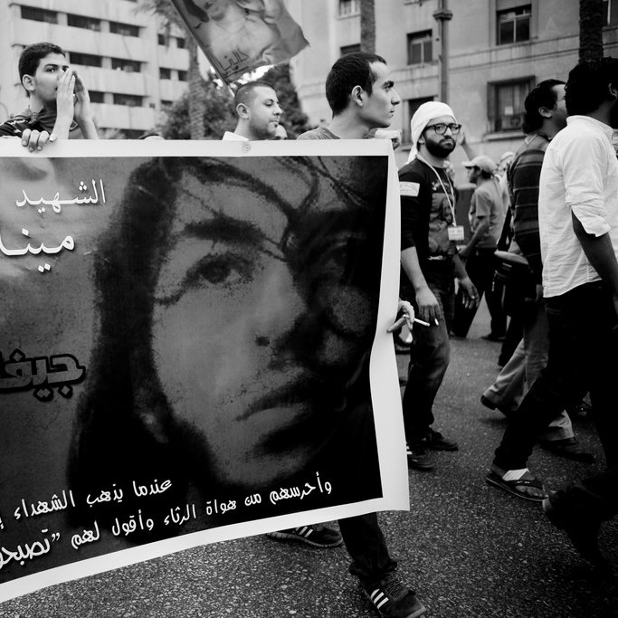 L'image montre une manifestation où des personnes portent une grande banderole. Sur la banderole, on peut voir le portrait d'une personne accompagnée d'un texte écrit en arabe, qui semble rendre hommage à cette personne. L'ambiance est chargée d'émotion, avec des manifestants en arrière-plan, et l'image est en noir et blanc, ce qui accentue la gravité de la situation. Les expressions des participants suggèrent une forte implication dans la cause défendue.