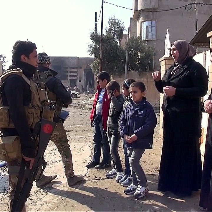 L'image montre une scène dans une rue dévastée, probablement dans une zone de conflit. Un soldat armé est debout, en uniforme militaire, tenant son arme. Il semble interagir avec deux femmes voilées qui lui parlent. À proximité, plusieurs enfants, dont deux garçons, regardent la scène. Le décor est marqué par des bâtiments endommagés et des débris qui témoignent d'une situation difficile. L'atmosphère semble tendue mais il y a aussi une interaction sociale entre les personnes présentes.