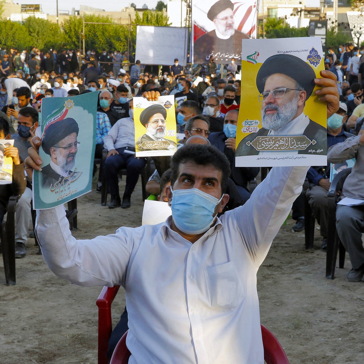 تظهر الصورة تجمعًا حاشدًا لمجموعة من الأشخاص يجلسون في مكان مفتوح. يحمل بعضهم لافتات أو صورًا لأحد الشخصيات السياسية. يرتدى الجميع تقريبًا كمامات واقية، مما يدل على الاحتراز من الأمراض. يبدو أن التجمع مرتبط بحدث سياسي أو دعم لشخص معين. الخلفية تضم أشجارًا ومباني، مما يعطي انطباعًا عن حدث في منطقة حضرية.