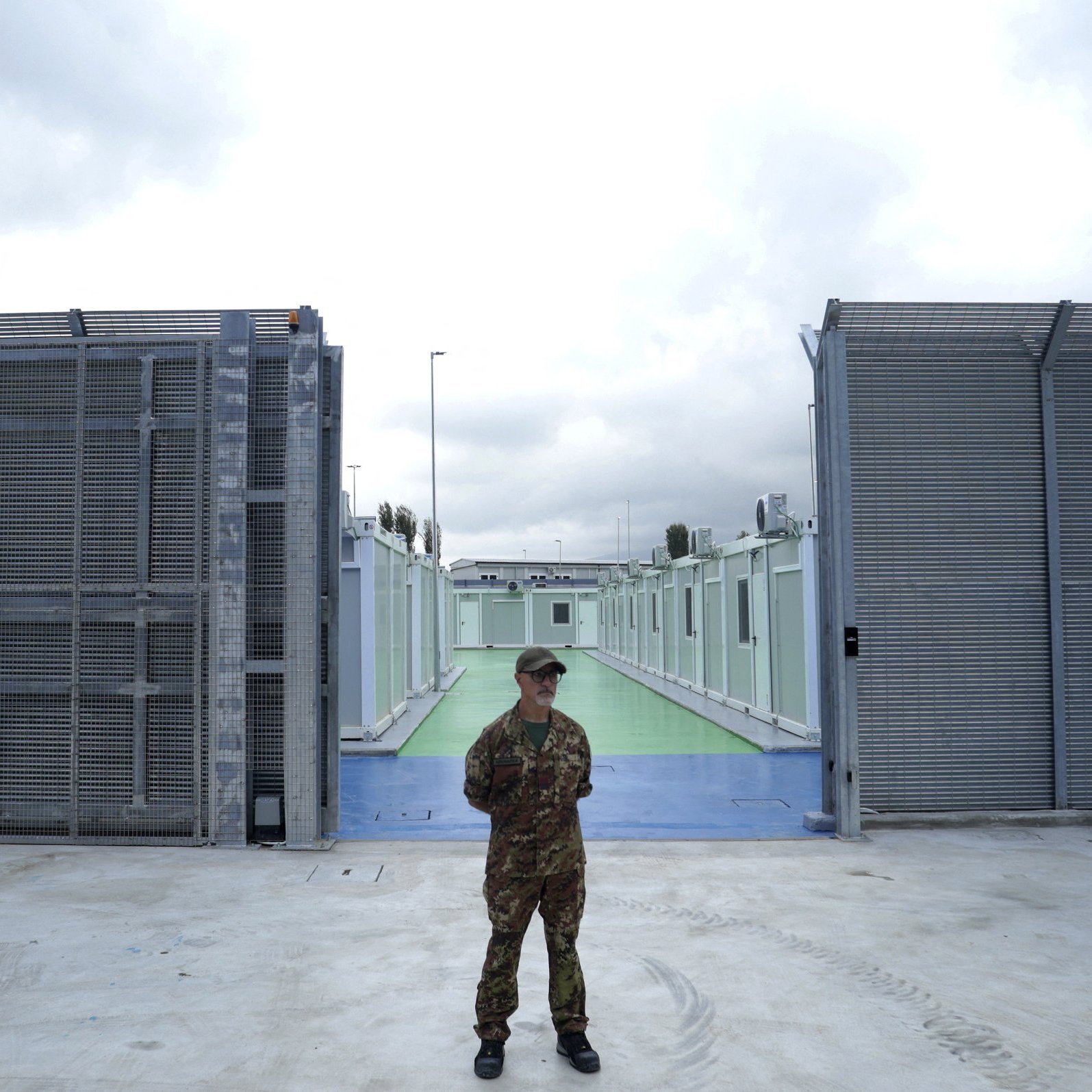 L'image montre un soldat en uniforme qui se tient au centre d'une zone sécurisée, probablement à l'extérieur d'une installation. À l'arrière-plan, on peut voir des structures métalliques grises et des murs, créant une ambiance de confinement. Le sol est en béton, et il y a une passerelle colorée qui semble être verte ou bleue. Le ciel est nuageux, suggérant un temps sombre. L'ensemble de l'image évoque un sentiment de sécurité renforcée et de surveillance.