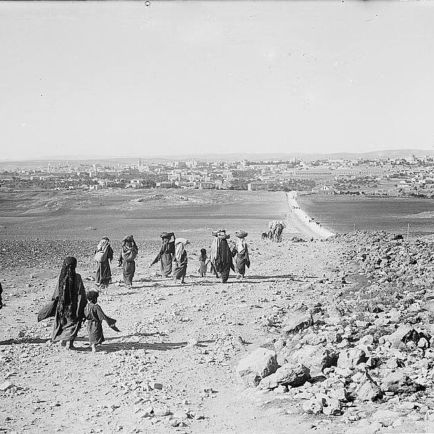 L'image montre un groupe de personnes marchant sur un chemin rural, entouré de paysages arides et de collines. Les individus, habillés de vêtements traditionnels, avancent ensemble, certains portant des charges sur la tête. En arrière-plan, on peut voir une ville avec des bâtiments, peut-être une petite agglomération, et une route qui serpente à travers le paysage. L'atmosphère semble sereine et la scène évoque un mode de vie traditionnel.