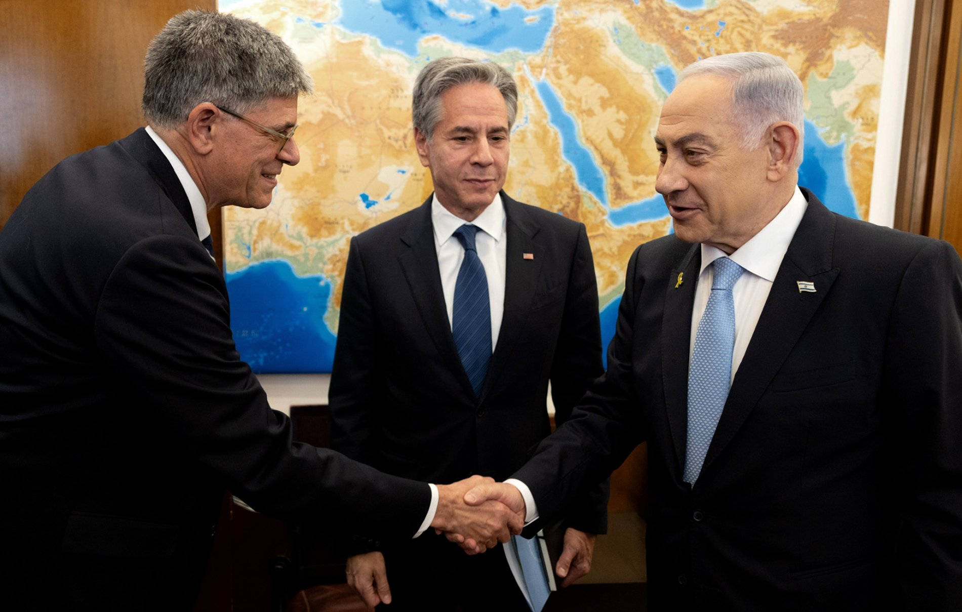 در تصویر، چند نفر در حال گپ‌وگفت هستند. دو نفر با هم دست می‌دهند و یکی دیگر در پس‌زمینه ایستاده است. پس‌زمینه شامل نقشه‌ای از منطقه خاورمیانه است. افراد در حال گفتگو به نظر می‌رسند و حالت چهره آن‌ها نشان‌دهنده تعامل مثبت است. لباس‌های رسمی آنها به اهمیت جلسه اشاره دارد.