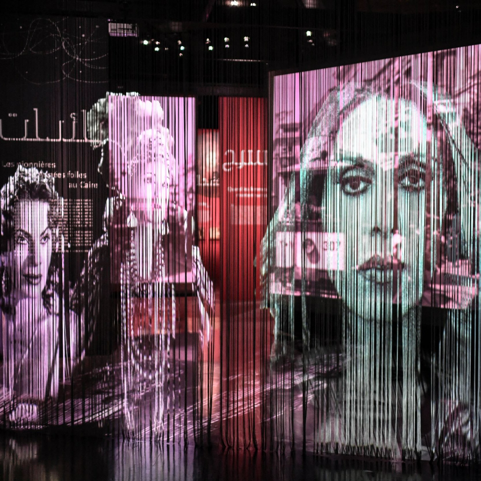 L'image présente une installation artistique composée de plusieurs grands panneaux transparents sur lesquels sont projetées des images en noir et blanc de visages féminins. Les projections créent un effet de profondeur et de mouvement, avec des formes et des silhouettes qui se superposent. Le fond est teinté de couleurs douces, suggérant une ambiance à la fois artistique et contemplative. Ce type d'installation évoque une réflexion sur la représentation et l'identité, tout en intégrant des éléments visuels captivants.