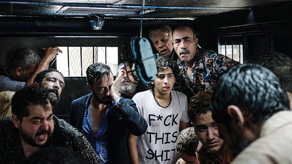 L'image montre un groupe de personnes dans un espace clos, visiblement en détresse ou en désespoir. Les visages expriment des émotions fortes, allant de la peur à l'angoisse. Certains sont habillés de manière décontractée, et l'un des individus porte un t-shirt avec un message provocateur. L'éclairage est faible, créant une ambiance sombre et oppressante. L'environnement suggère une situation de tension ou de crise.