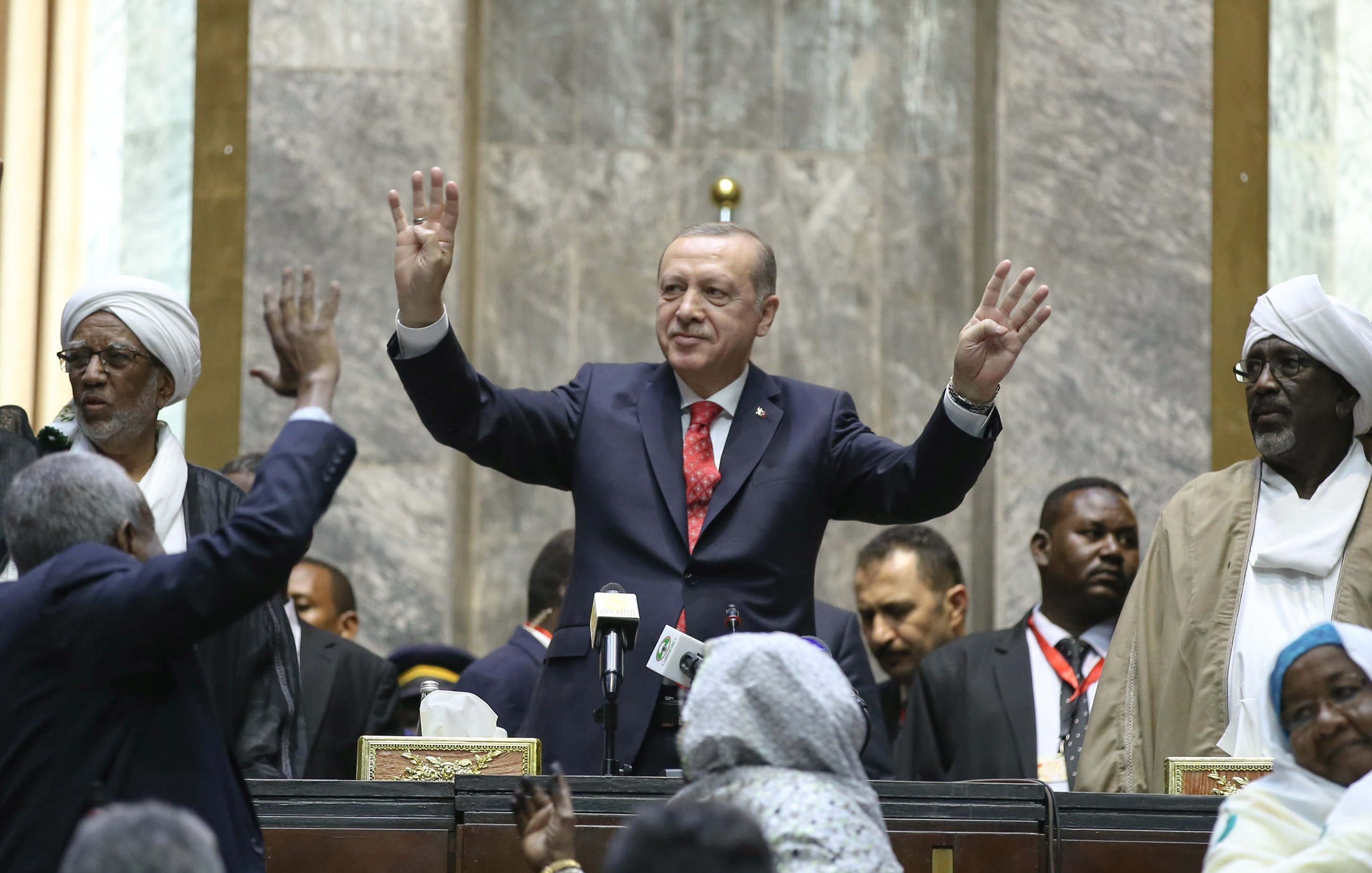 L'image montre un homme debout au centre, levant les mains en signe de salutation ou de triomphe. Il est entouré d'une foule de personnes qui semblent célébrer ou applaudir. Le décor suggère un cadre formel, probablement une assemblée ou un événement officiel, avec des personnes en habits traditionnels et des caméras visibles en arrière-plan. L'atmosphère semble être celle d'une occasion importante, marquée par une interaction entre le orateur et le public.