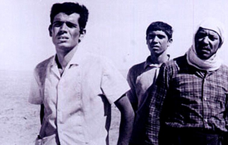 L'image représente trois hommes se tenant dans un paysage désertique. Leurs expressions faciales semblent refléter l'inquiétude ou la tristesse. Le premier homme, à gauche, a les traits anguleux et semble regarder au loin. Le deuxième, au centre, paraît plus jeune et affiche une expression de curiosité ou de perplexité. Le dernier homme, à droite, porte un foulard autour de la tête et a une apparence plus âgée, ce qui pourrait suggérer qu'il a vécu des épreuves. Le fond du paysage est vaste et vide, ce qui accentue l'impression d'isolement et de dureté.