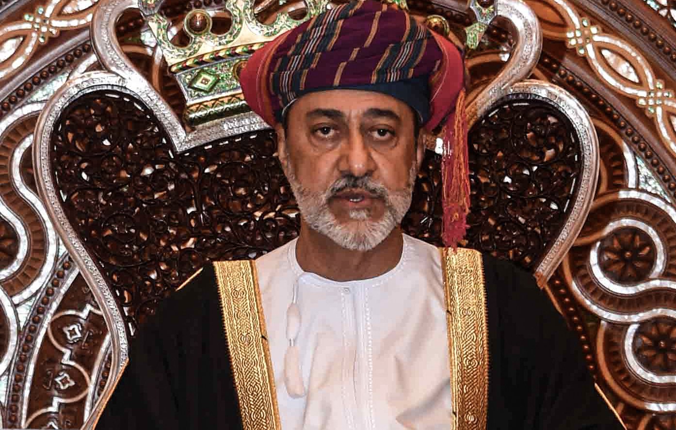 L'image montre un homme assis sur un trône orné. Il porte un costume traditionnel, comprenant une robe blanche et un turban coloré. Son expression est sérieuse et solennelle. Le fond est richement décoré avec des motifs complexes, créant une ambiance majestueuse. Les détails de la couronne et du trône mettent en valeur son statut élevé.