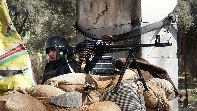 L'image montre un soldat en arrière-plan, vêtu d'un uniforme et portant un casque. Devant lui, il y a une mitrailleuse montée sur un trépied, posée sur un tas de sacs de sable. Le contexte semble être une zone de conflit, avec des éléments de construction en arrière-plan, probablement dans une région touchée par des combats. La scène évoque des mesures de protection et de défense.