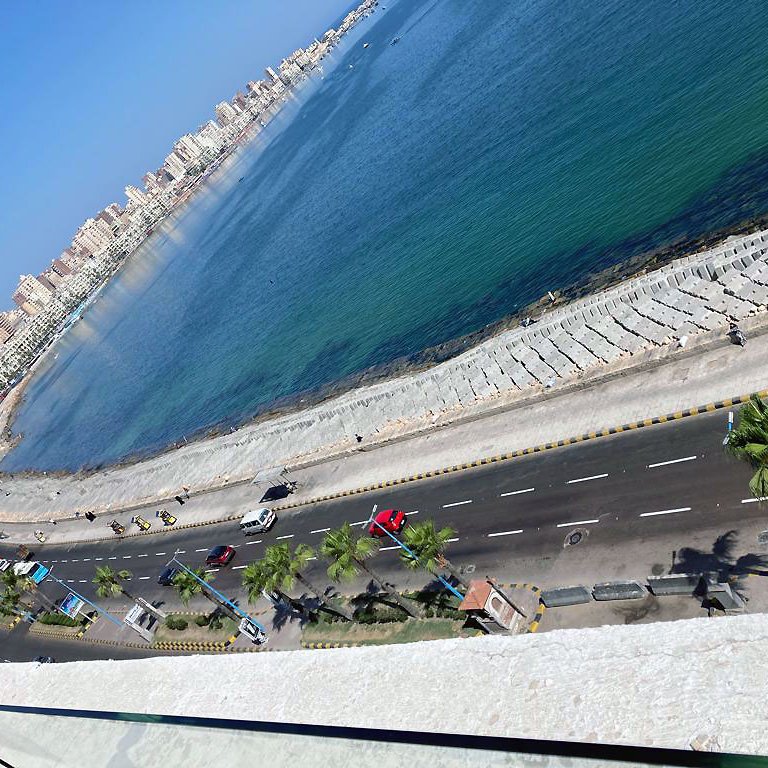 L'image montre une vue panoramique d'une côte en bord de mer. On aperçoit une route qui longe la plage, avec des palmiers sur le côté. L'eau se présente dans des teintes de bleu et de vert, et on peut voir des bâtiments en fond, probablement la ville ou des complexes résidentiels. La lumière du soleil illumine la scène, donnant une atmosphère agréable et ensoleillée.