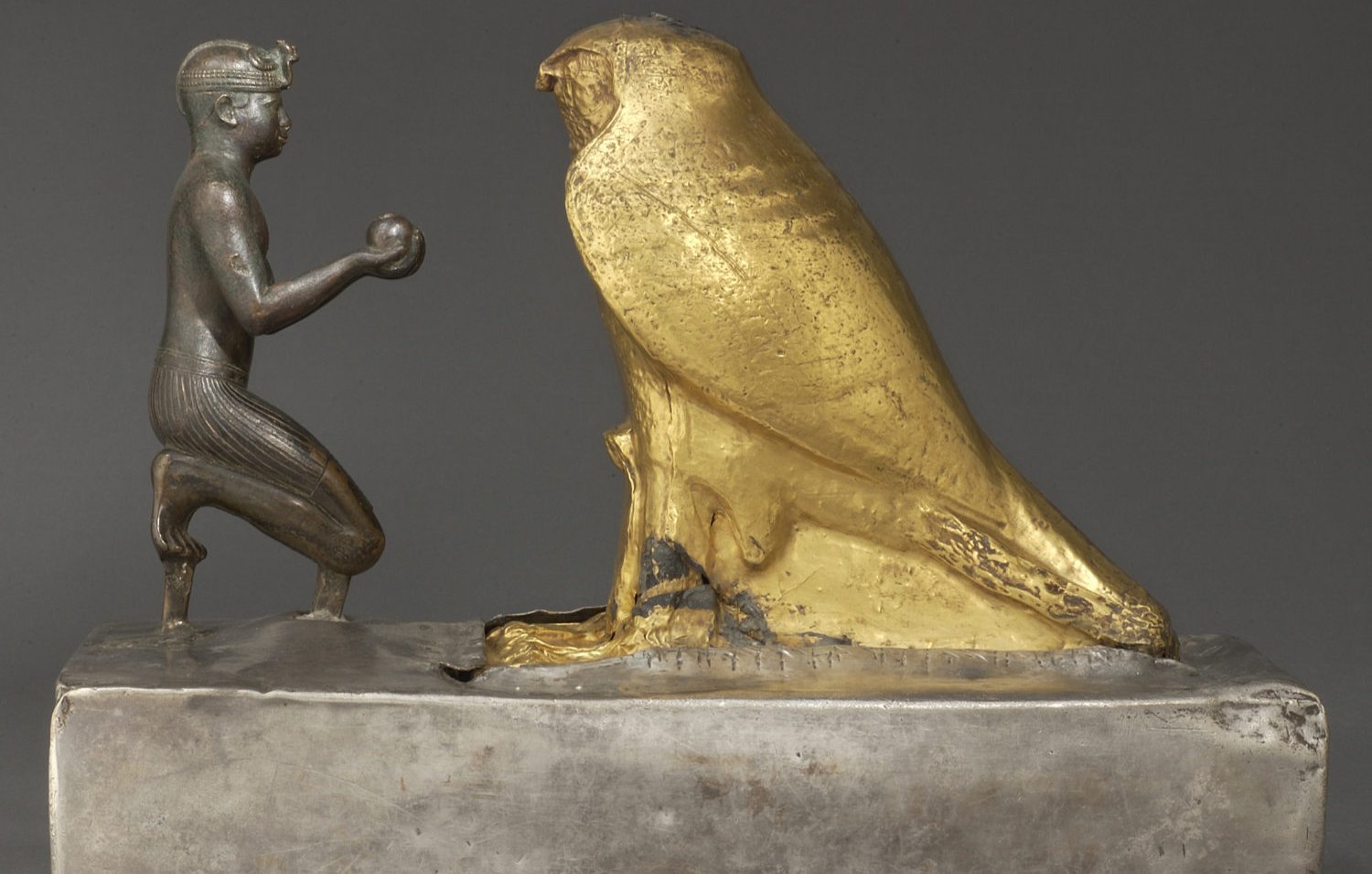 L'image représente une sculpture en métal, où l'on peut voir un personnage humain à genoux tenant un objet dans ses mains, face à un grand hibou doré. Le personnage est sculpté de manière détaillée, avec des traits qui évoquent un style ancien, probablement lié à l'Égypte ou à une culture similaire. Le hibou, de couleur dorée, symbolise peut-être la sagesse ou un aspect divin. La base de la sculpture est en métal argenté, offrant un contraste intéressant avec la brillance du hibou doré. Cette œuvre semble revêtir une signification symbolique ou religieuse.