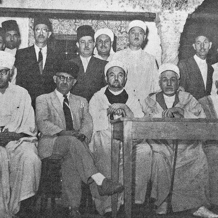 L'image montre un groupe de personnes rassemblées dans une pièce. Elles portent des vêtements traditionnels, notamment des turbans et des robes longues. Au centre, quelques individus sont assis à une table, tandis que d'autres se tiennent debout derrière eux. L'atmosphère semble formelle et solennelle, suggérant une réunion ou une cérémonie. Les murs sont en pierre, ce qui donne un aspect historique à l'ensemble.