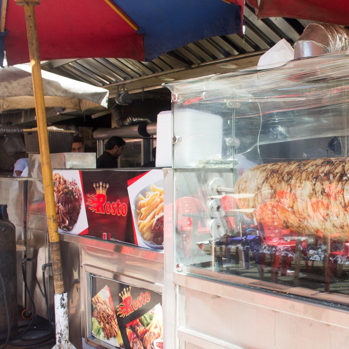 The image depicts a food stall featuring a large kebab or shawarma that's roasting on a vertical spit. The meat is visibly browning as it cooks, and there are flames underneath. The stall is covered by colorful umbrellas providing shade. In the foreground, a man stands behind the counter, likely the vendor, appearing to interact with customers or prepare food. The stall also displays menu items on signs, emphasizing its offerings. The atmosphere looks lively, typical of a street food setting.
