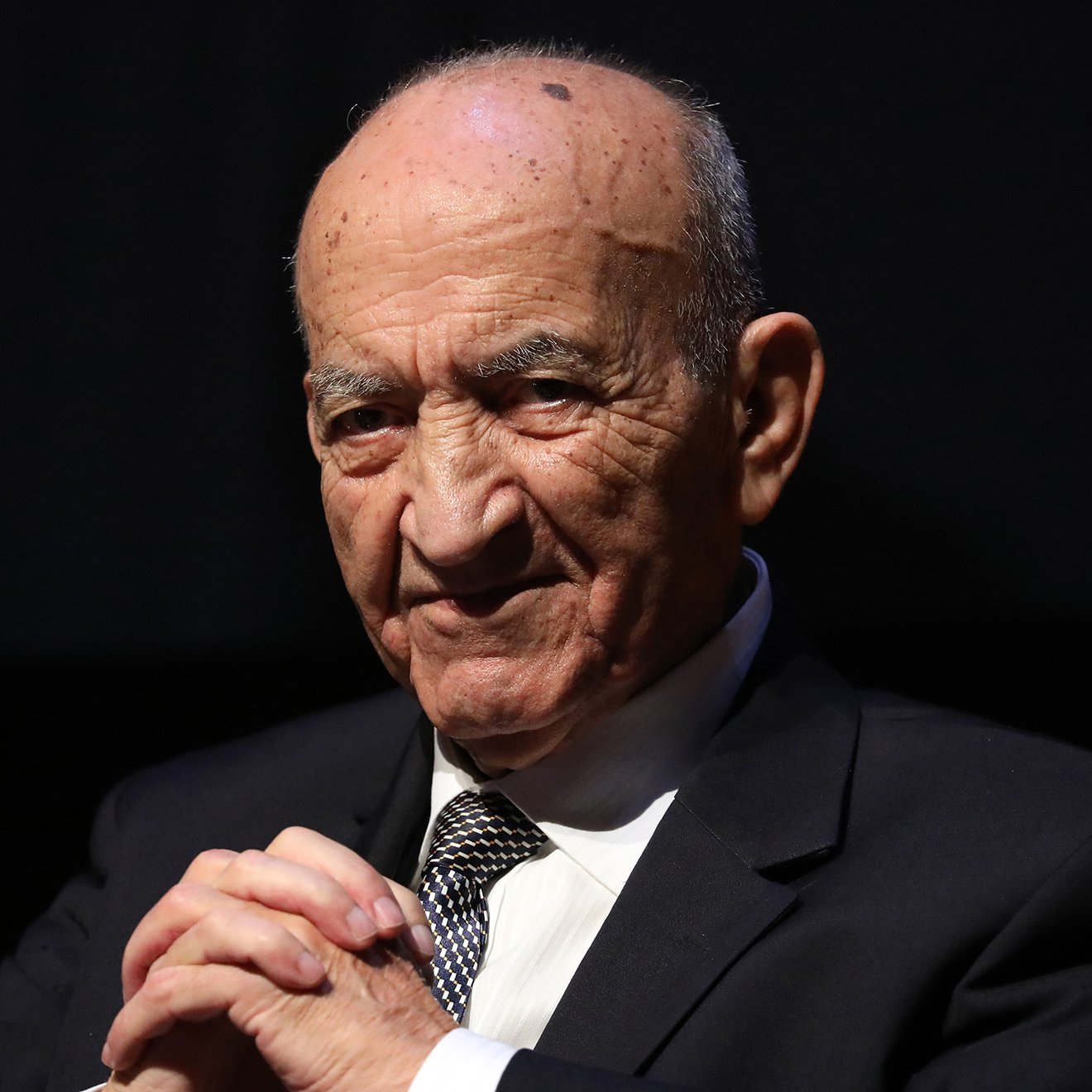 L'image montre un homme âgé, portant un costume noir et une cravate rayée. Il a une expression sérieuse sur son visage, avec un léger sourire. Ses mains sont posées l'une sur l'autre devant lui, et il semble être en train de réfléchir ou d'écouter attentivement. Le fond est sombre, ce qui met en valeur son visage et sa tenue.