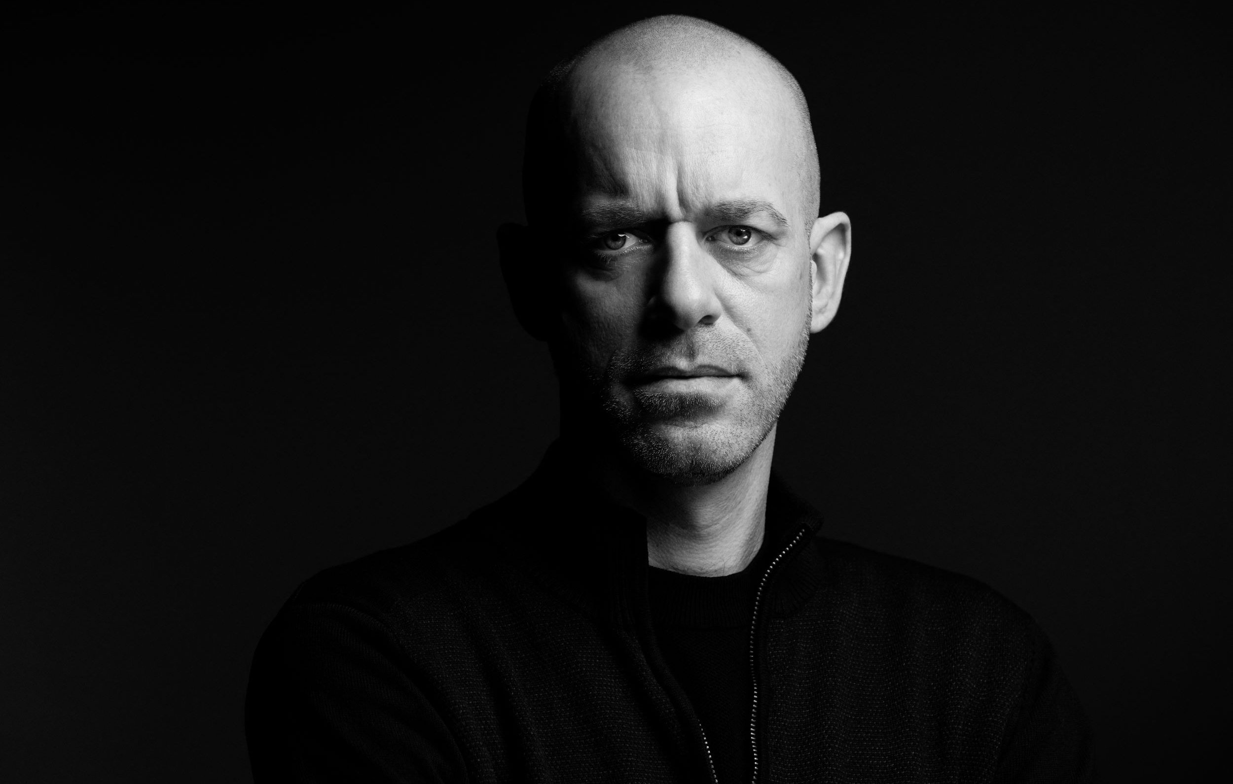 الصورة تُظهر رجلًا ذو رأس أصلع، يقف في ضوء خافت مما يبرز ملامح وجهه بوضوح. عيونه تحمل تعبيرًا جادًا وقويًا، بينما يحدق بشكل مباشر نحو الكاميرا. يرتدي سترة داكنة، ويبدو أن الخلفية سوداء، مما يضيف إلى جمالية الصورة ويعطي إحساسًا بالدراماتيكية.