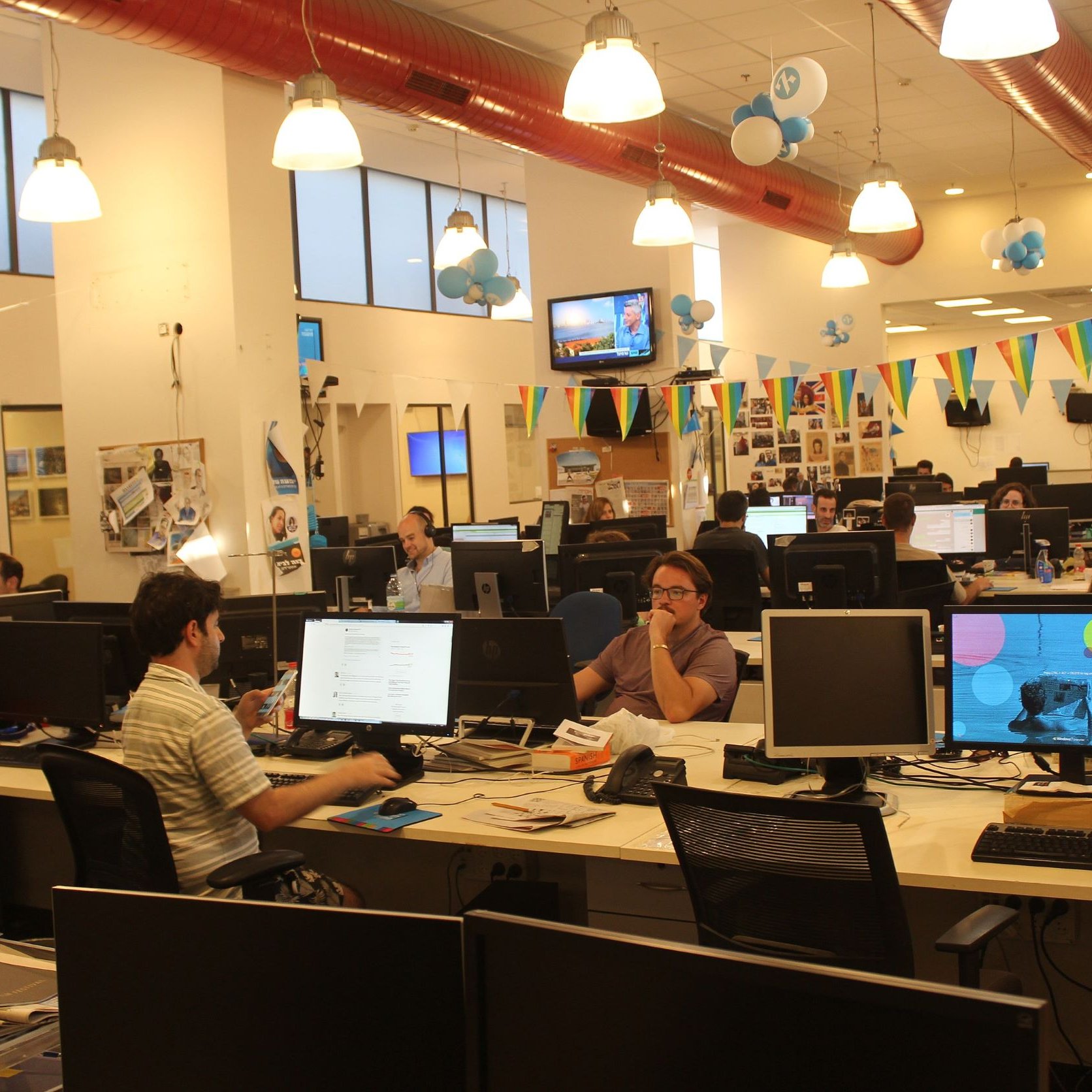 The image shows an open office space with several workstations. There are multiple desks equipped with computers, where people are engaged in various tasks. The decor includes colorful balloons and banners hanging from the ceiling, suggesting a festive atmosphere. Large windows allow for ample natural light, and the overall ambiance seems lively and collaborative, with employees working both individually and in small groups.