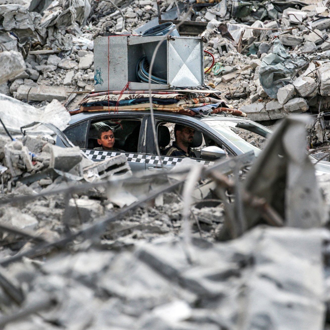 L'image montre un paysage dévasté avec des décombres sur le sol. Au centre, on aperçoit une voiture dans laquelle se trouvent deux personnes. La voiture semble être coincée parmi les débris, qui comprennent des morceaux de béton et d'autres matériaux d'une structure effondrée. L'ambiance générale dégage une impression de destruction et de chaos, témoignant d'une situation difficile.