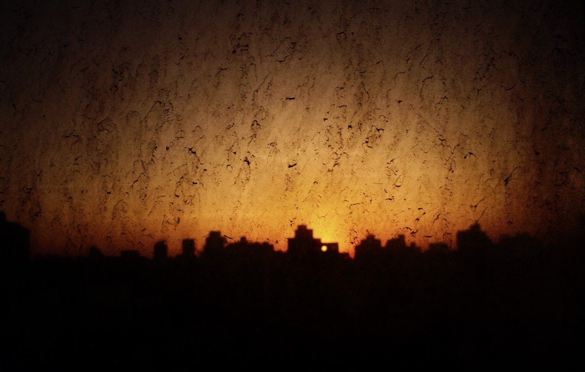 L'image montre un paysage urbain au crépuscule, avec des silhouettes de bâtiments se découpant contre un ciel orangé. La texture de la vitre est visible, ajoutant une dimension bruitée et floue à la scène. Les reflets et les imperfections de la vitre créent une atmosphère légèrement mystérieuse, tandis que la lumière du soleil couchant rappelle la fin de la journée.