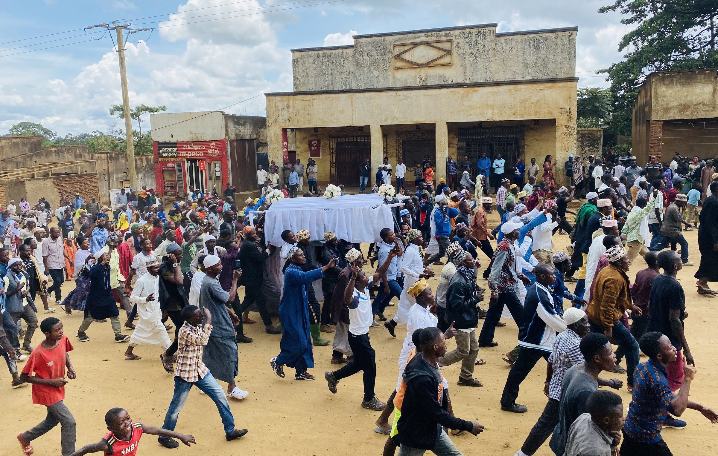 L'image montre une grande foule de personnes marchant dans une rue, probablement lors d'un événement ou d'une procession. Au centre, il y a un chariot couvert d'un drap blanc, et les gens semblent marcher avec une certaine ferveur. On peut apercevoir différents styles vestimentaires, suggérant une diversité parmi les participants. En arrière-plan, des bâtiments sont visibles, ainsi qu'un ciel partiellement nuageux. L'atmosphère semble collective et déterminée.