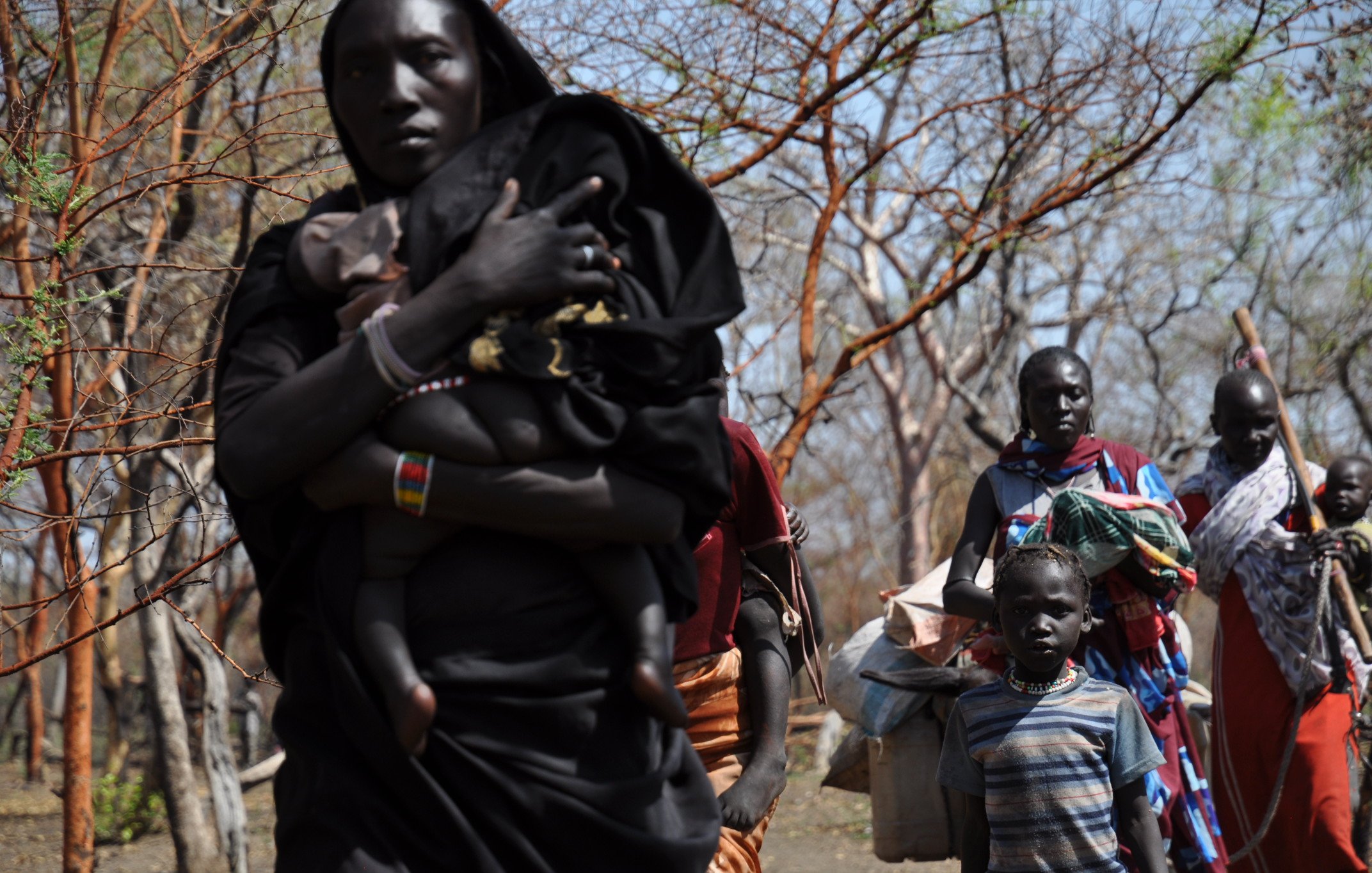 L'image montre un groupe de personnes se déplaçant dans une zone boisée. Au premier plan, une femme porte un enfant sur son dos, tandis qu'à côté d'elle, un petit garçon regarde vers l'avant. D'autres femmes et enfants les suivent, portant des sacs et des objets sur leurs épaules. Leurs vêtements sont colorés, et le décor est constitué d'arbres aux branches dénudées, suggérant un environnement aride. L'atmosphère semble être celle d'un déplacement ou d'une communauté en mouvement.