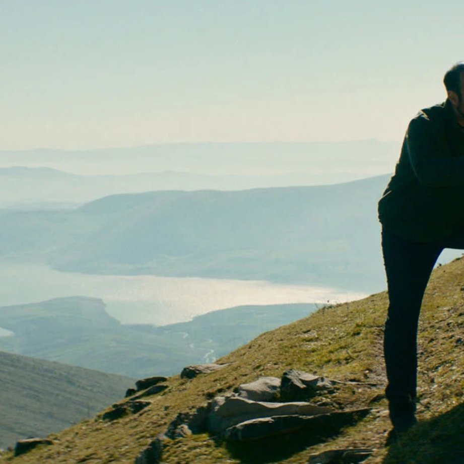 L'image montre un homme et un enfant sur une colline, face à un paysage naturel vaste et montagneux. L'homme semble parler à l'enfant, peut-être pour lui donner des conseils ou partager un moment d'intimité. En arrière-plan, on aperçoit un lac scintillant et des montagnes, le tout baigné par une lumière douce, ce qui crée une atmosphère sereine et contemplative. La scène évoque des thèmes de connexion et de découverte en pleine nature.