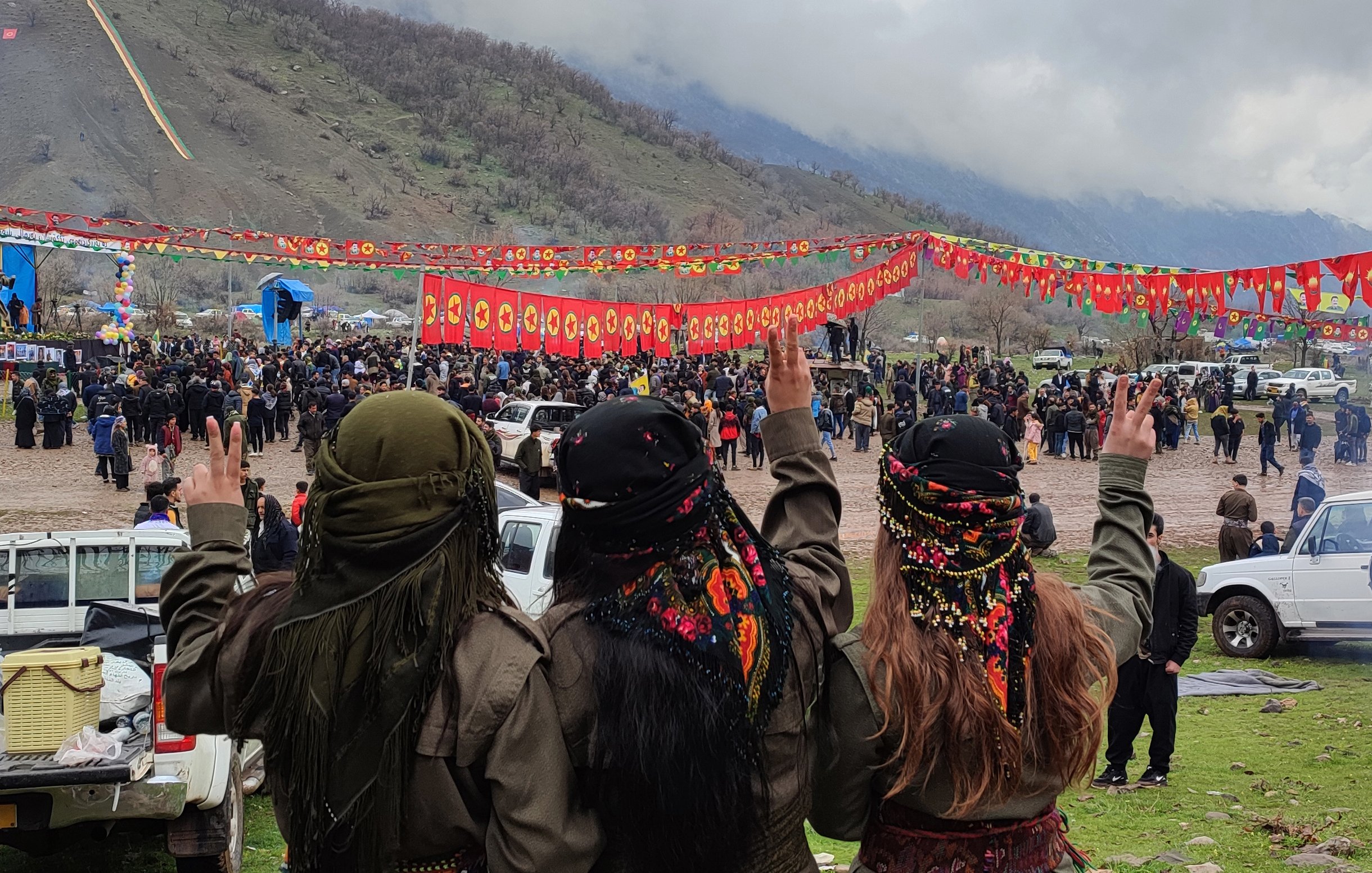 L'image montre un groupe de trois personnes vues de dos, faisant un signe de paix avec leurs mains. Elles portent des vêtements traditionnels et des foulards colorés. En arrière-plan, on peut voir une foule rassemblée pour un événement, avec des banderoles rouges décorées de symboles. Le paysage environnant est montagnard et sous un ciel nuageux. L'atmosphère semble festive et solennelle.