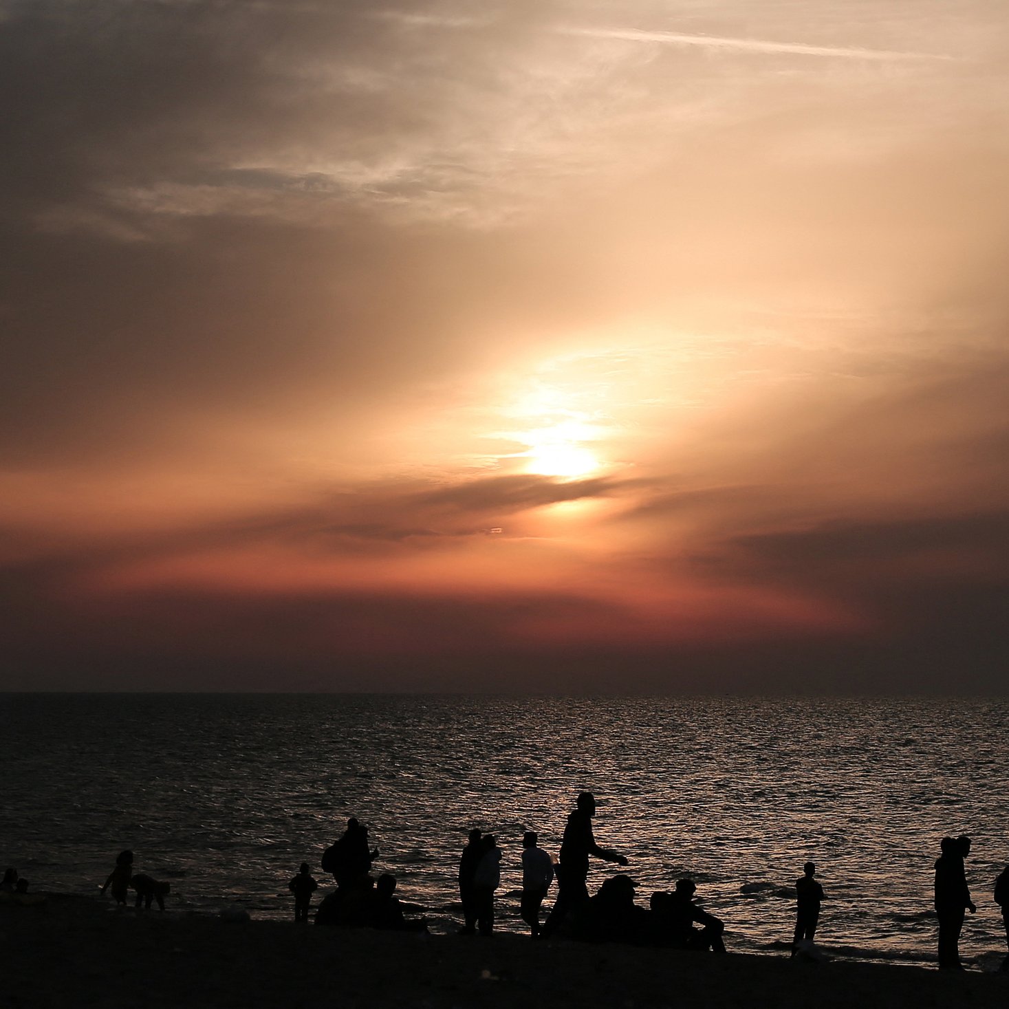L'immagine mostra una scena al tramonto sulla spiaggia. Il cielo è dipinto di tonalità calde, con sfumature di arancione e rosa, mentre il sole si avvicina all'orizzonte. In primo piano, si vedono diverse silhouette di persone che camminano lungo la riva, alcune sedute o accoccolate sulla sabbia. La superficie dell'acqua riflette i colori del cielo, creando un'atmosfera calma e suggestiva.