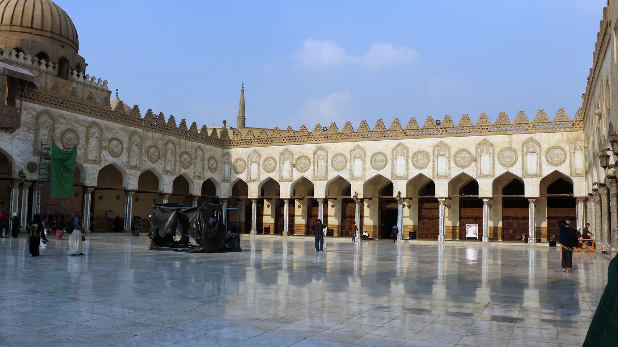 الصورة تظهر ساحة كبيرة داخل معلم تاريخي، يحتمل أن يكون مسجدًا أو جامعة إسلامية. الساحة محاطة بأقواس زخرفية وبهياكل عالية تظهر الهندسة المعمارية الإسلامية التقليدية. الأرض مغطاة ببلاط لامع يعكس ضوء السماء، مما يضفي جمالية خاصة على المكان. في الخلفية، يمكن رؤية قبة وجدران مزخرفة، بالإضافة إلى بعض الأشخاص الذين يتجولون في المكان. السماء زرقاء مع بعض الغيوم، مما يضيف لمسة من الهدوء إلى المشهد.