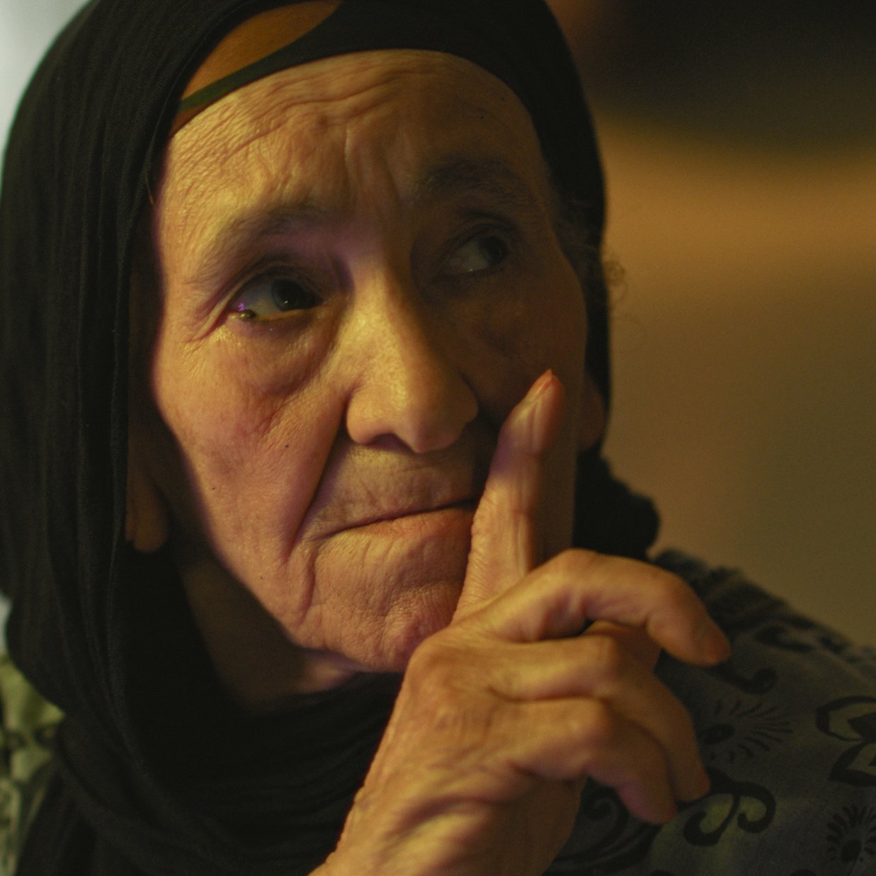 L'image montre une femme âgée avec un regard pensif. Elle porte un foulard noir qui encadre son visage. Son expression est sérieuse et elle semble faire un geste de silence avec son doigt posé sur ses lèvres. La lumière dans le fond est douce, créant une ambiance calme et introspective. Les détails de son visage reflètent une certaine sagesse et vécu.