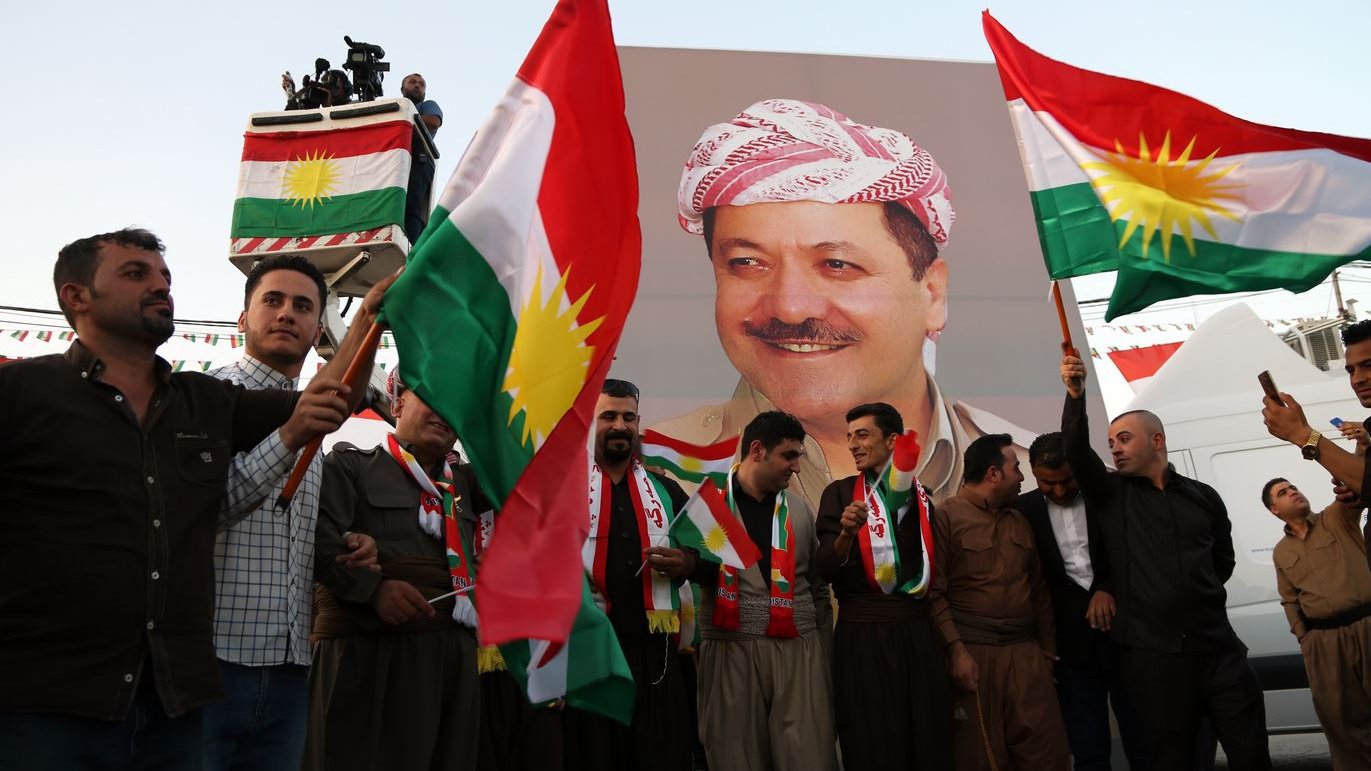 در تصویر گروهی از مردان لباس کردی به همراه پرچم‌های کردستان را مشاهده می‌کنیم. آن‌ها در حال ایستادن در مقابل یک بنر بزرگ هستند که تصویر فردی را نمایش می‌دهد. افراد با نشاط و خوشحالی پرچم‌ها را در دست دارند و به نظر می‌رسد که در یک مراسم یا جشن خاص شرکت کرده‌اند. فضای جمعی و پر انرژی به خوبی در تصویر حس می‌شود.