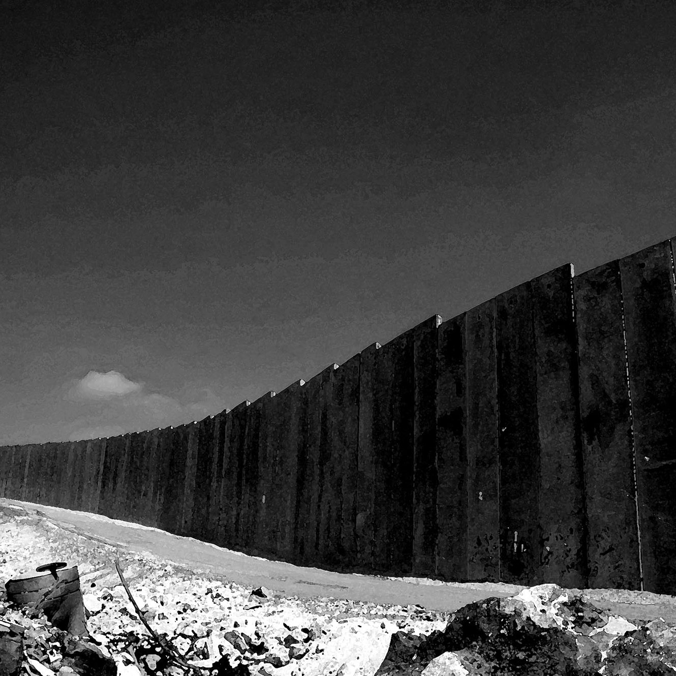 الصورة تظهر جدارًا مرتفعًا ومتينًا يمتد على طول الأفق، مما يوحي بعزلة أو تفرقة. الأرض المحيطة بالجدار تبدو غير مستوية وتحتوي على صخور وحطام، مما يعكس بيئة قاسية. السماء في الخلفية تبدو صافية ولكنها تتناقض مع قساوة الجدار. الصورة بألوان الأسود والأبيض، مما يضيف طابعًا دراميًا وحزينًا للمشهد.