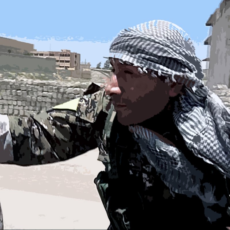 L'image montre un homme en tenue militaire, portant un foulard sur la tête. Il semble concentré et se trouve près d'un baril. En arrière-plan, on aperçoit des bâtiments et un paysage urbain. La scène évoque une ambiance de stress ou de tension, caractéristique des zones de conflit.