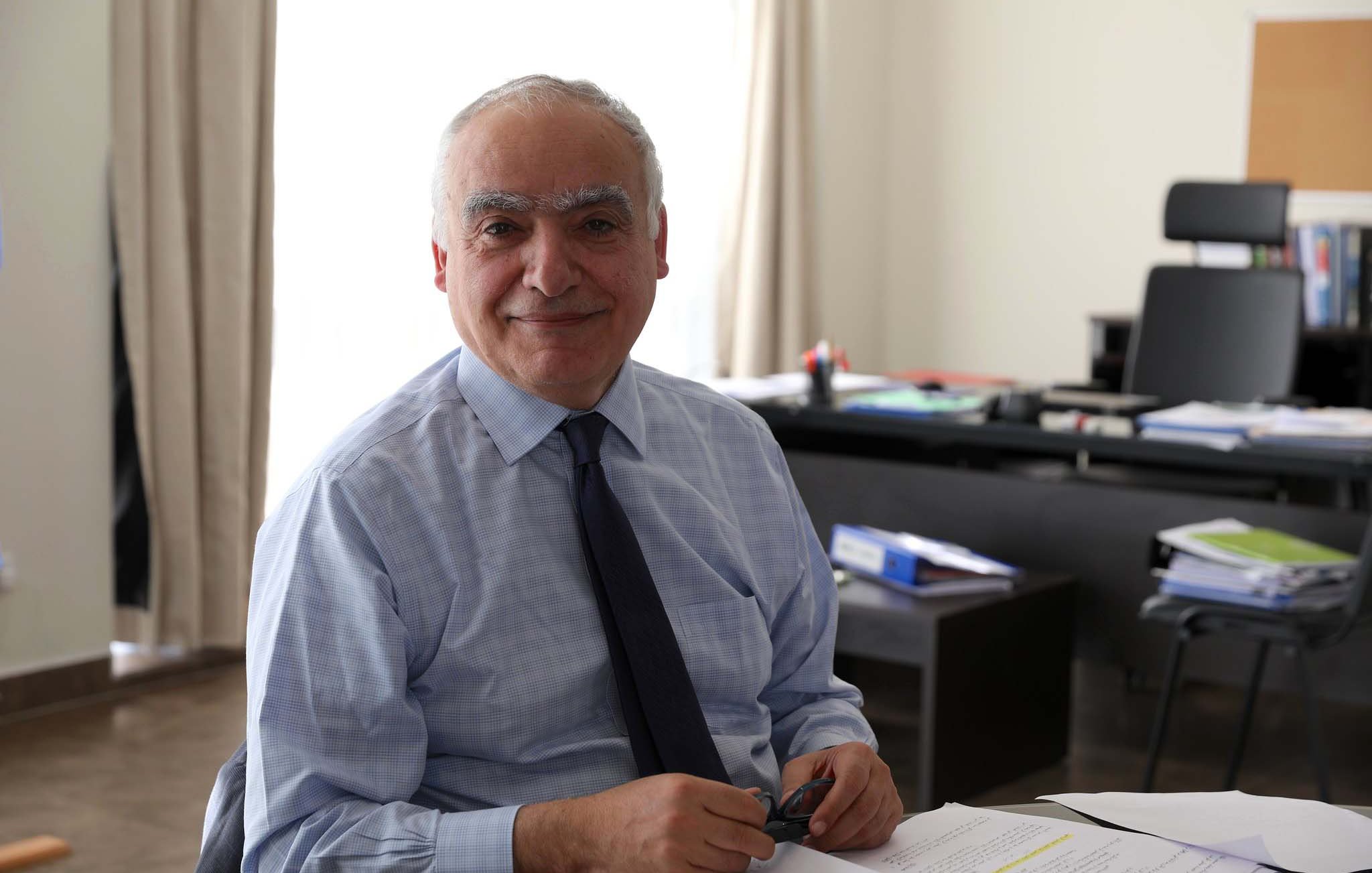 در این تصویر، مردی با موهای خاکستری و لبخند نرم نشسته است. او پیراهن آبی کمرنگ و یک کراوات تیره به تن دارد. در پس‌زمینه، یک میز کار با تعدادی اسناد و فایل‌ها دیده می‌شود. نور طبیعی از پنجره‌های بزرگ به داخل می‌تابد و فضایی روشن و دوستانه ایجاد کرده است. فضای اتاق به طور کلی ساده و مدرن به نظر می‌رسد.