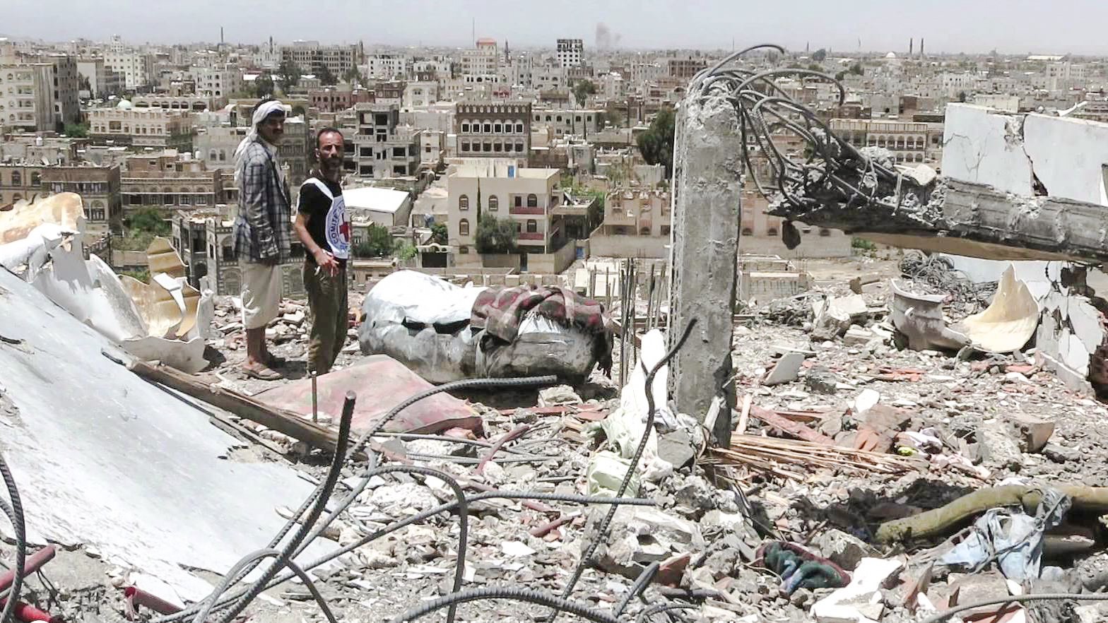 L'image montre deux hommes se tenant au milieu des décombres d'un bâtiment détruit, surplombant une ville en arrière-plan. Les structures environnantes semblent endommagées, avec de nombreux bâtiments partiellement effondrés et une apparence de désolation. Le ciel est clair, mais l'environnement porte des traces évidentes de conflit. Les hommes semblent examine la scène, témoignant d'une situation difficile et dévastatrice.