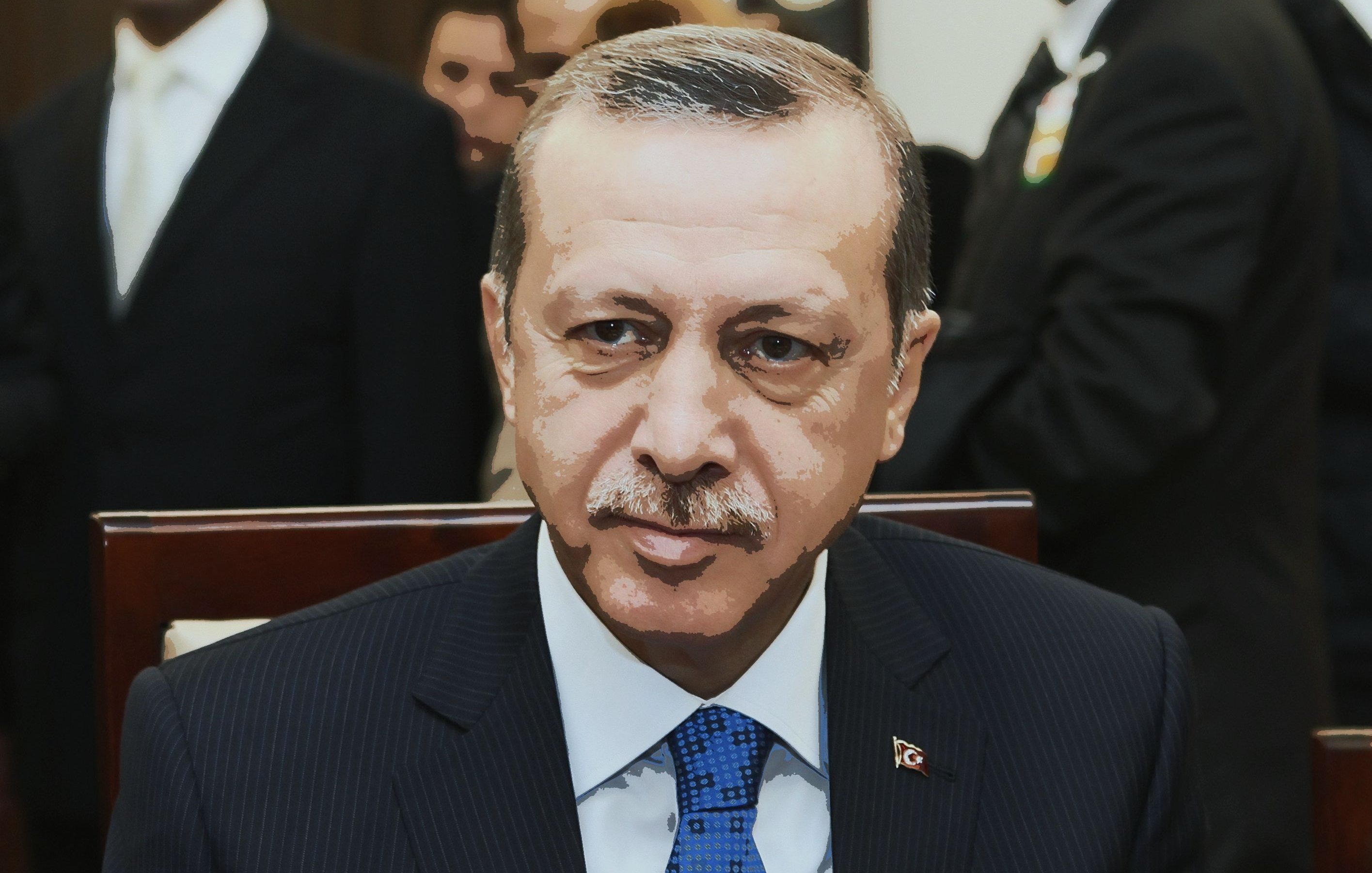 Je ne sais pas qui est cette personne, mais l'image montre un homme en costume noir avec une cravate bleue. Il a une expression sérieuse et semble être dans un cadre formel avec d'autres personnes en arrière-plan.