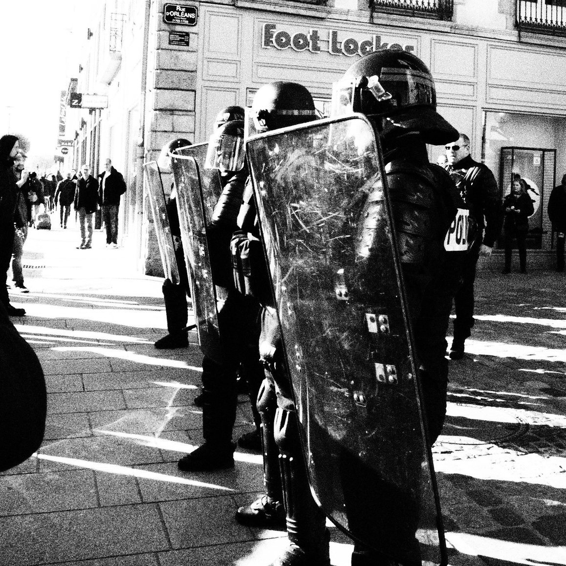 L'image montre une scène urbaine en noir et blanc, où des policiers en tenue anti-émeute, portant des casques et tenant des boucliers, se tiennent en position sur une rue. Ils sont face à une foule de personnes, dont certaines semblent prendre des photos. L'environnement est composé de bâtiments commerciaux, et l'ambiance semble tendue. Les ombres et la lumière créent un contraste saisissant dans la composition.