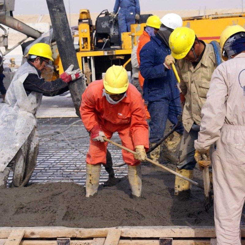 L'image montre plusieurs ouvriers en train de travailler sur un chantier de construction. Ils portent des casques de sécurité jaunes et des vêtements de protection, tels que des combinaisons et des bottes en caoutchouc. L'un des travailleurs utilise une pelle pour manipuler du béton, tandis que d'autres semblent s'occuper de l'installation ou de la préparation du béton. En arrière-plan, on peut voir des machines de chantier et des équipements. L'atmosphère est active, indiquant un environnement de travail intense.