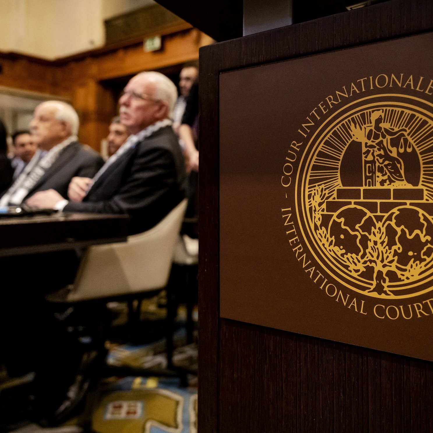 L'image montre une scène à la Cour internationale de justice. Au premier plan, on peut voir le symbole de la Cour, qui est un emblème distinctif. En arrière-plan, plusieurs personnes sont assises, probablement des membres du tribunal ou des délégués, attendant une session ou un discours. L'environnement semble formel, avec une décoration typique d'une institution judiciaire.