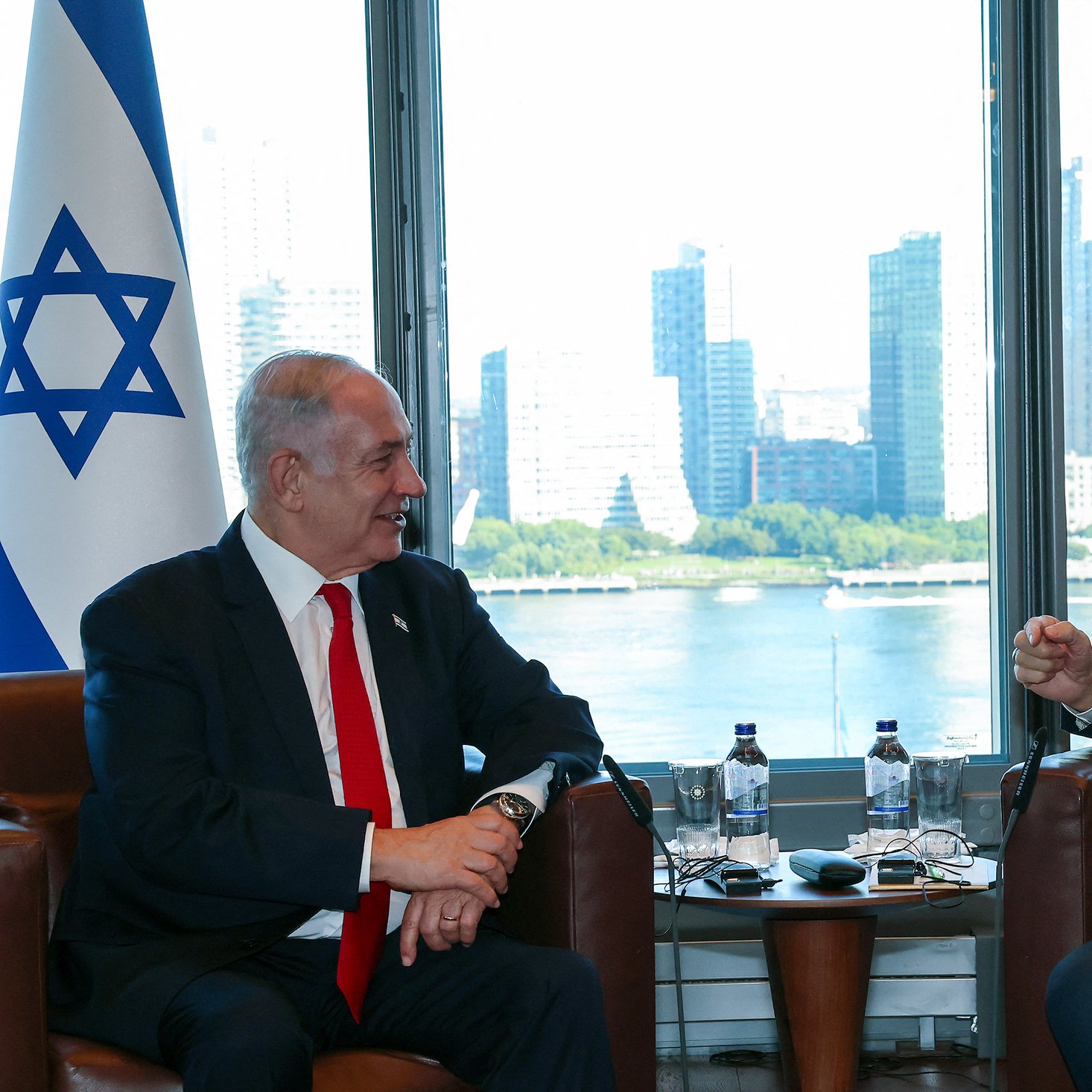 این تصویر دو فرد را در حال گفت‌وگو نشان می‌دهد. در سمت چپ، پرچم اسرائیل و در سمت راست، پرچم ترکیه قرار دارد. این افراد در یک اتاق با نمایی از شهر در پس‌زمینه نشسته‌اند که ساختمان‌های بلند و دریاچه‌ای را نشان می‌دهد. هر دو نفر به نظر می‌رسد که در حال تبادل نظر دوستانه هستند. 