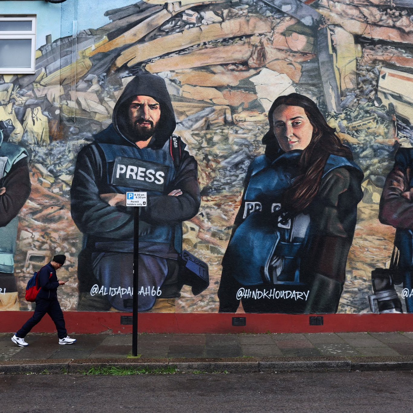 L'immagine mostra un murale che ritrae quattro giornalisti con abbigliamento di protezione e giubbotti con la scritta "PRESS". Sono posizionati in modo prominente davanti a un contesto di distruzione, simile a un'area colpita da un terremoto. Una persona sta camminando lungo il marciapiede di fronte al murale. I volti dei giornalisti esprimono determinazione e serietà, sottolineando il loro lavoro e l'importanza della libertà di stampa.