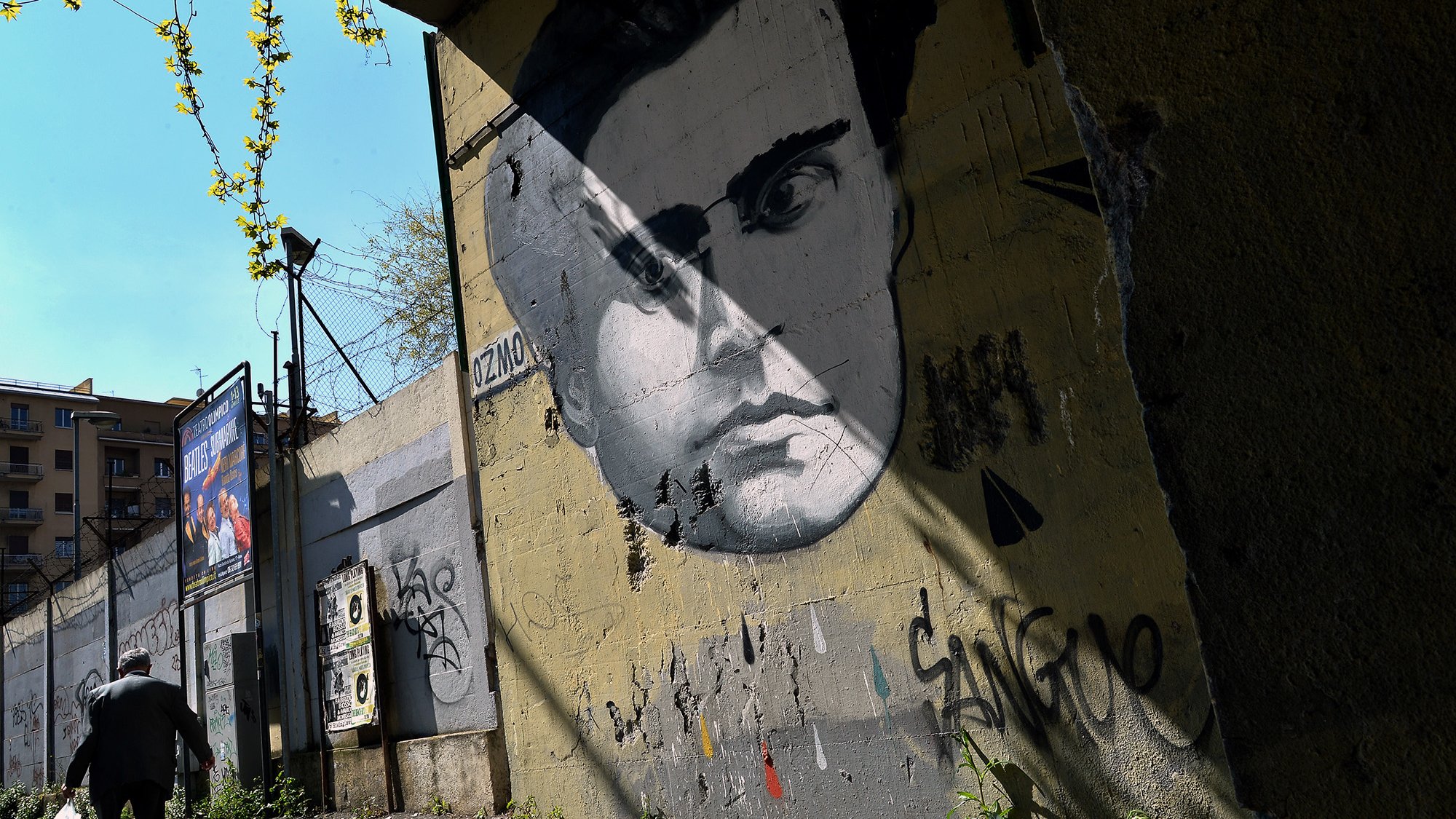 L'immagine mostra un murale di un volto umano, molto probabilmente di un uomo, dipinto su un muro grigio. Il viso ha lineamenti distintivi e intensi, con un'espressione che attira l'attenzione. Sullo sfondo, si può vedere una strada o un marciapiede con un uomo che cammina, mentre alcuni elementi di verde e graffiti decorano la scena. La luce del sole crea una serie di ombre, accentuando l'atmosfera urbana del luogo.