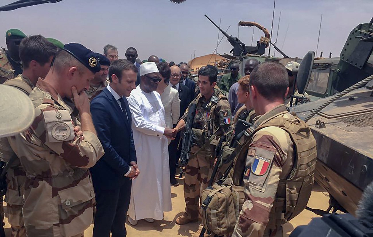 تظهر الصورة مجموعة من الجنود والعسكريين في منطقة صحراوية. في الوسط، يوجد رجل يرتدي لباسًا تقليديًا أبيض، بصحبة شخصية أخرى يبدو أنها رسمية. العسكريون يرتدون زيًا عسكريًا ويحملون الأسلحة، بينما يظهر في الخلفية بعض المركبات العسكرية. الأجواء تبدو جدية، والمشهد يوحي بلقاء رسمي أو زيارة تفقدية.