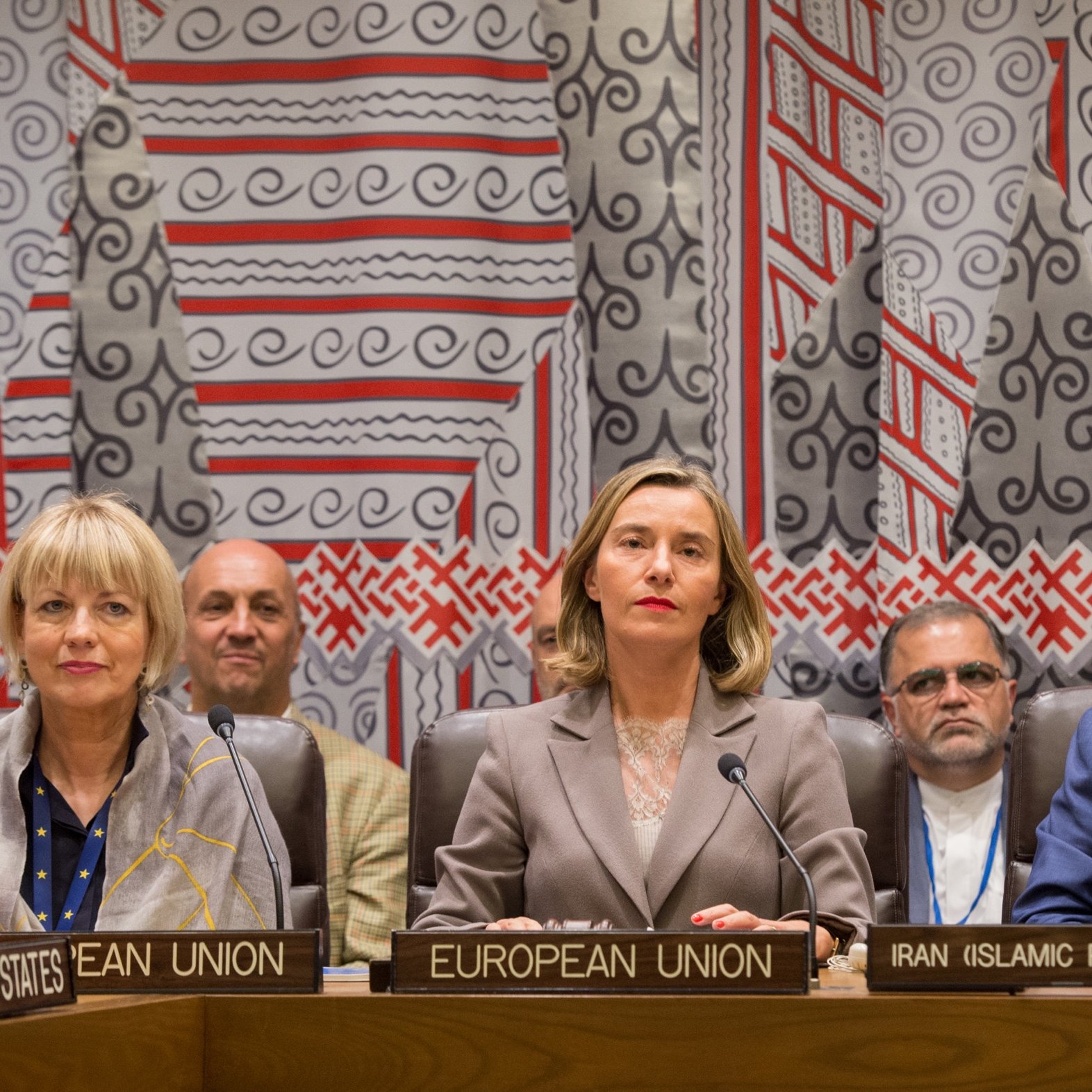 در این تصویر، تعدادی از افراد در جلسه‌ای رسمی نشسته‌اند. در ردیف جلو، افراد نشسته بر روی صندلی‌هایی با برچسب‌های مشخص شامل "اتحادیه اروپا"، "ایران (جمهوری اسلامی)" و "ایالات متحده" هستند. زنانی که در وسط نشسته است، در لباس رسمی به نظر می‌رسد و توجه بیشتری به خود جلب می‌کند. پس‌زمینه تصویر با الگوهای خاصی تزئین شده که فضای رسمی و دیپلماتیک را رقم می‌زند. افراد دیگر در تصاویر به وضوح دیده می‌شوند و در حالت تمرکز به میزگرد توجه دارند.