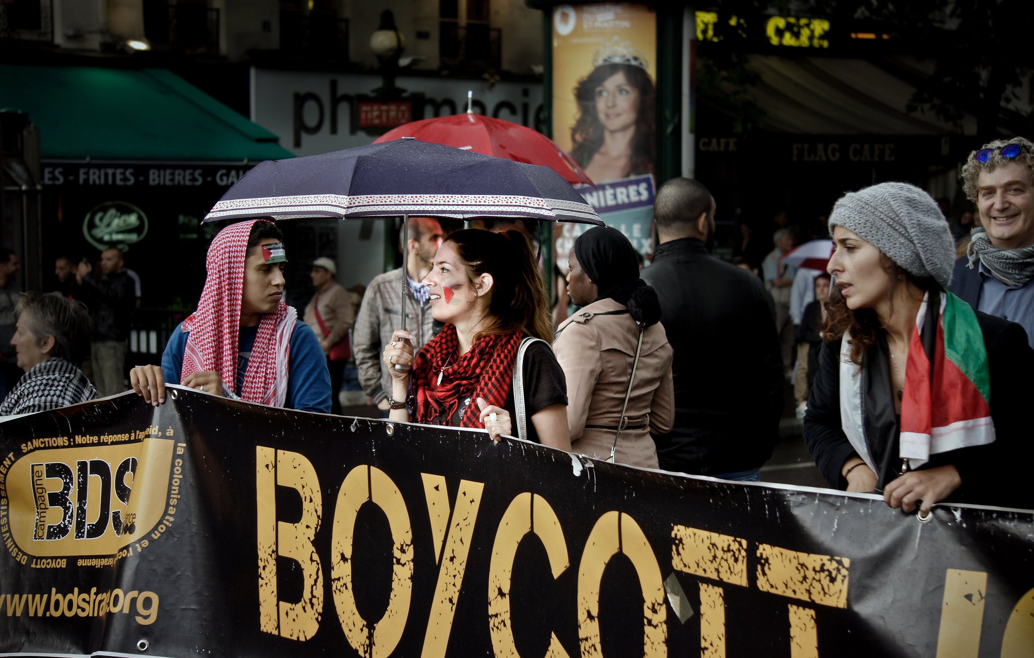 L'image montre un groupe de personnes participant à une manifestation. Au premier plan, on voit une femme tenant un grand panneau qui dit "BOYCOTT". Elle est entourée de plusieurs manifestants, dont certains portent des accessoires ou des vêtements aux couleurs de différents pays. Il y a aussi des parapluies, probablement à cause de la météo. L'ambiance semble engagée, avec un mélange de détermination et de solidarité parmi les participants.