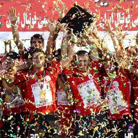 L'image montre une équipe de football célébrant une victoire. Les membres de l'équipe portent des maillots rouges et affichent des sourires de joie. Ils tiennent des trophées et sont entourés de confettis colorés qui tombent sur eux. L'ambiance est festive, et on peut sentir l'excitation de la victoire à travers leurs expressions. Des banderoles et des affiches derrière eux montrent le nom de l'équipe et le mot "champion".