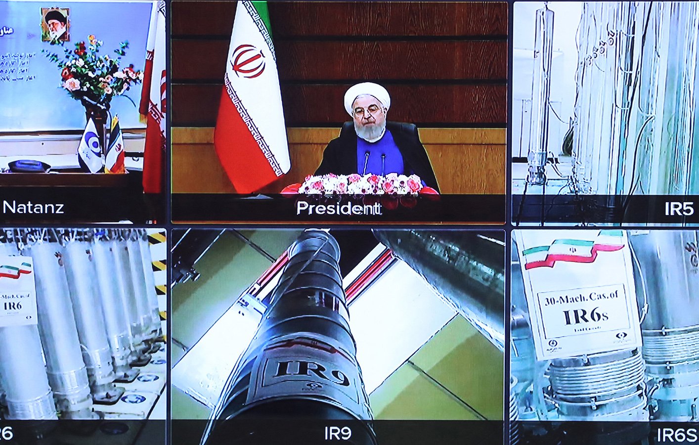 L'image montre une composition de plusieurs écrans de télévision. Au centre, on voit un homme, probablement un président, assis devant un bureau avec des fleurs, entouré de drapeaux iraniens. Sur les autres écrans, il y a des images d'installations ou d'équipements liés à la technologie nucléaire, avec des tubes et des étiquettes indiquant différentes séries de centrifugeuses nucléaires. Il y a aussi des symboles et des drapeaux qui décrivent les différentes installations mentionnées. L'ensemble semble se rapporter à un discours ou une présentation sur le programme nucléaire de l'Iran.