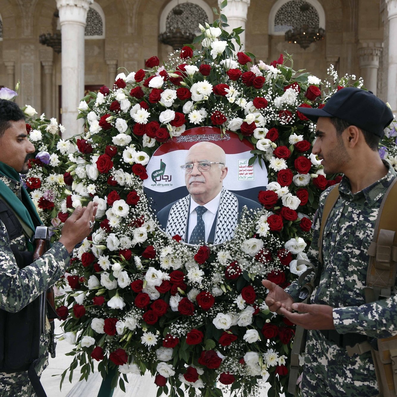 Deux hommes en uniforme discutent devant une couronne de fleurs avec un portrait au centre.