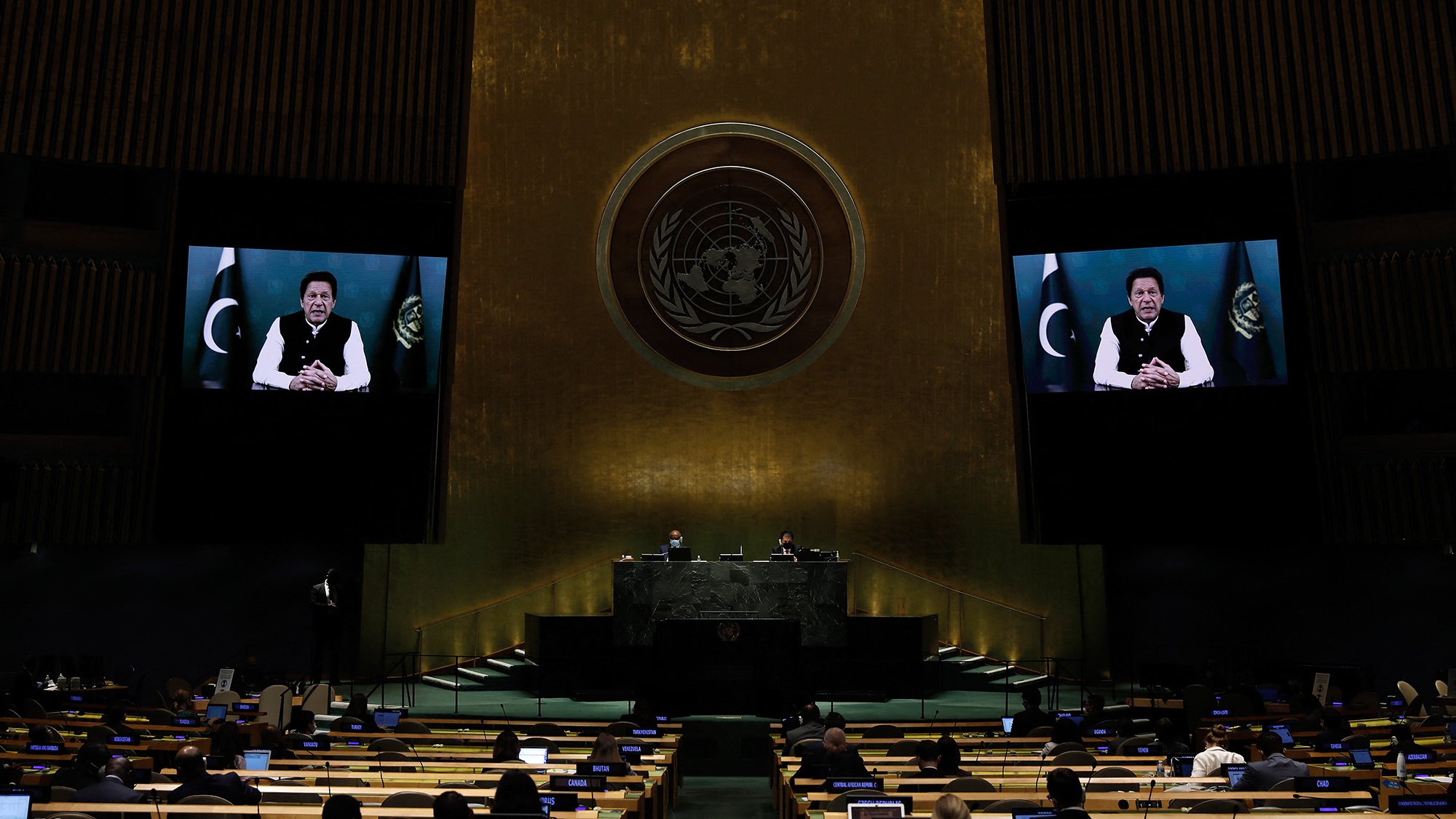 L'image montre une scène à l'Assemblée générale des Nations Unies. On peut voir des délégués assis à des bureaux, prêts à participer à la réunion. Un grand écran affiche une personne qui s'adresse à l'assemblée, tandis que d'autres officiels sont visibles sur la scène principale derrière le pupitre. L'arrière-plan est décoré avec l'emblème des Nations Unies, symbolisant l'importance de cet organe international. L'atmosphère est formelle et solennelle.
