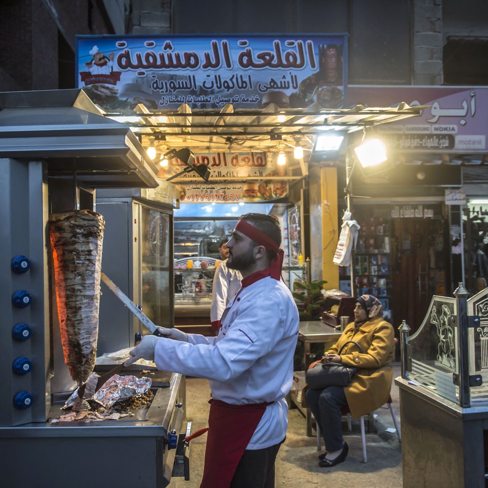 Sur cette image, on peut voir un vendeur de nourriture dans une rue animée, probablement dans un pays du Moyen-Orient. Il est en train de préparer un plat cuisiné, et on peut apercevoir un grand rouleau de viande, typique des stands de kebab ou de shawarma. Le vendeur porte une tenue de chef, composée d'une veste blanche et d'un couvre-chef. À côté de lui, une autre personne semble attendre, assise et observant. L'ambiance est chaleureuse, avec des lumières attirantes au-dessus du stand et des panneaux indiquant le nom du lieu.