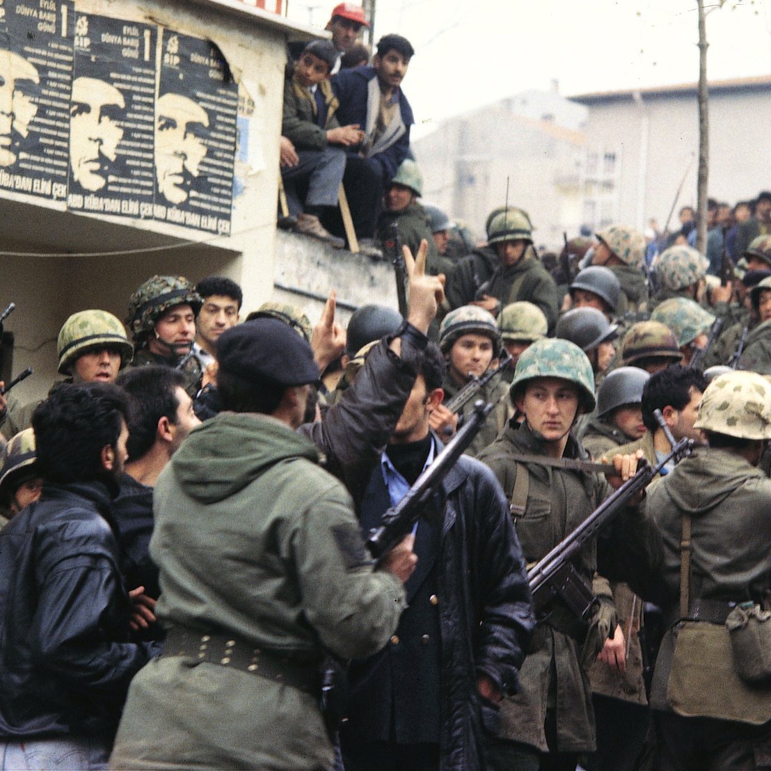 L'image montre un groupe de soldats en uniforme, portant des casques et armés, réunis autour d'une foule. On peut voir des hommes en civil, certains semblent s'opposer aux militaires. En arrière-plan, des affiches sont visibles sur le mur, peut-être représentant des figures politiques ou des messages de protestation. L'atmosphère est tendue et indique un moment de conflit ou de manifestation. Les visages des individus expriment des émotions variées, allant de la détermination à l'inquiétude.