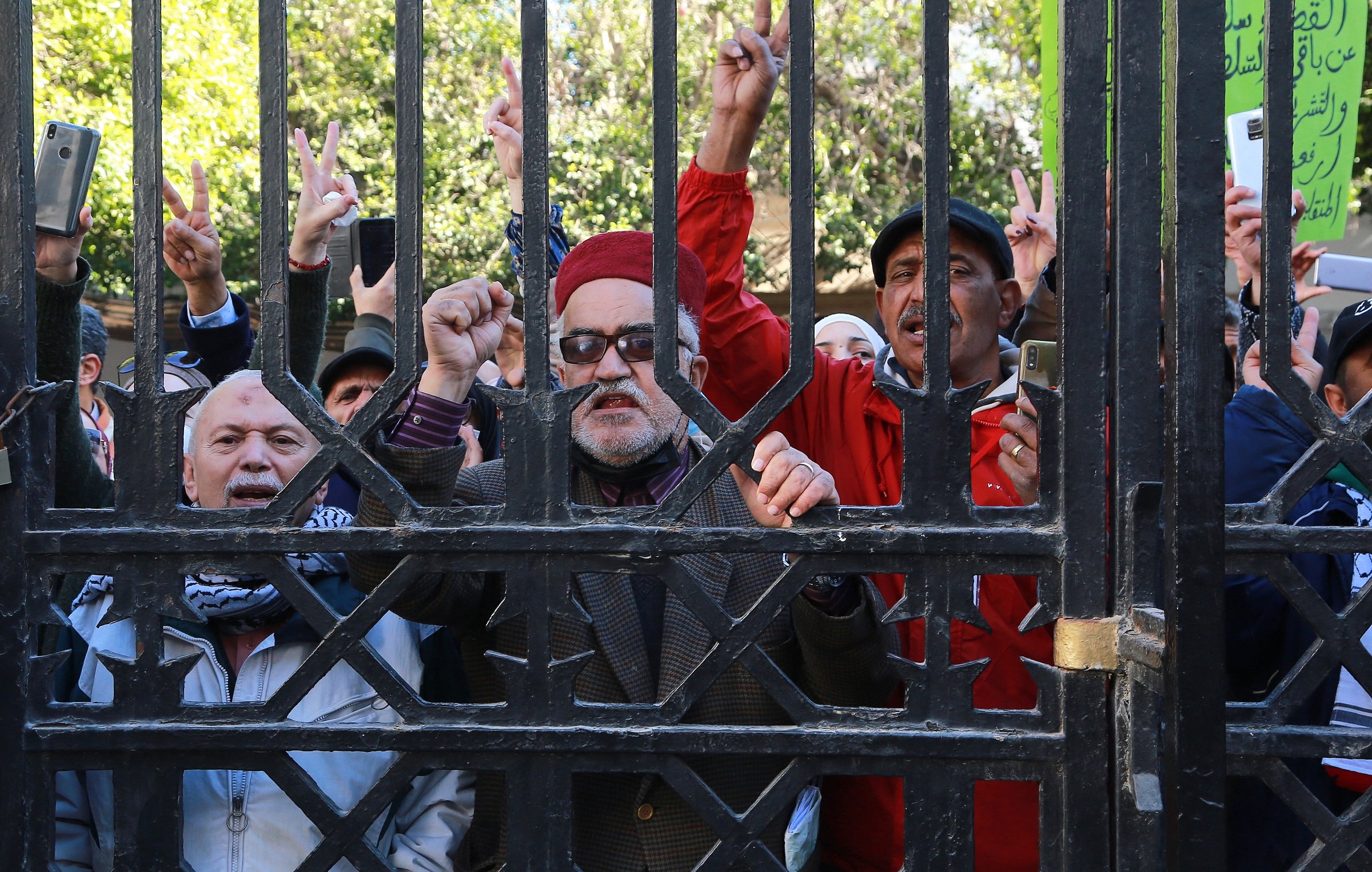 L'image montre un groupe de personnes rassemblées derrière une grille. Certains individus lèvent leurs doigts en forme de V, symbole de la paix ou de la victoire. Les expressions des participants varient, allant de la détermination à l'enthousiasme. Il semble qu'ils soient engagés dans une manifestation ou un rassemblement. Les arrière-plans sont flous, mettant l'accent sur les manifestants et leur activité.
